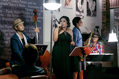 A musical performance featuring a female vocalist on stage with a jazz band, including a double bassist, saxophonist, and pianist, in a cozy venue with ambient lighting.