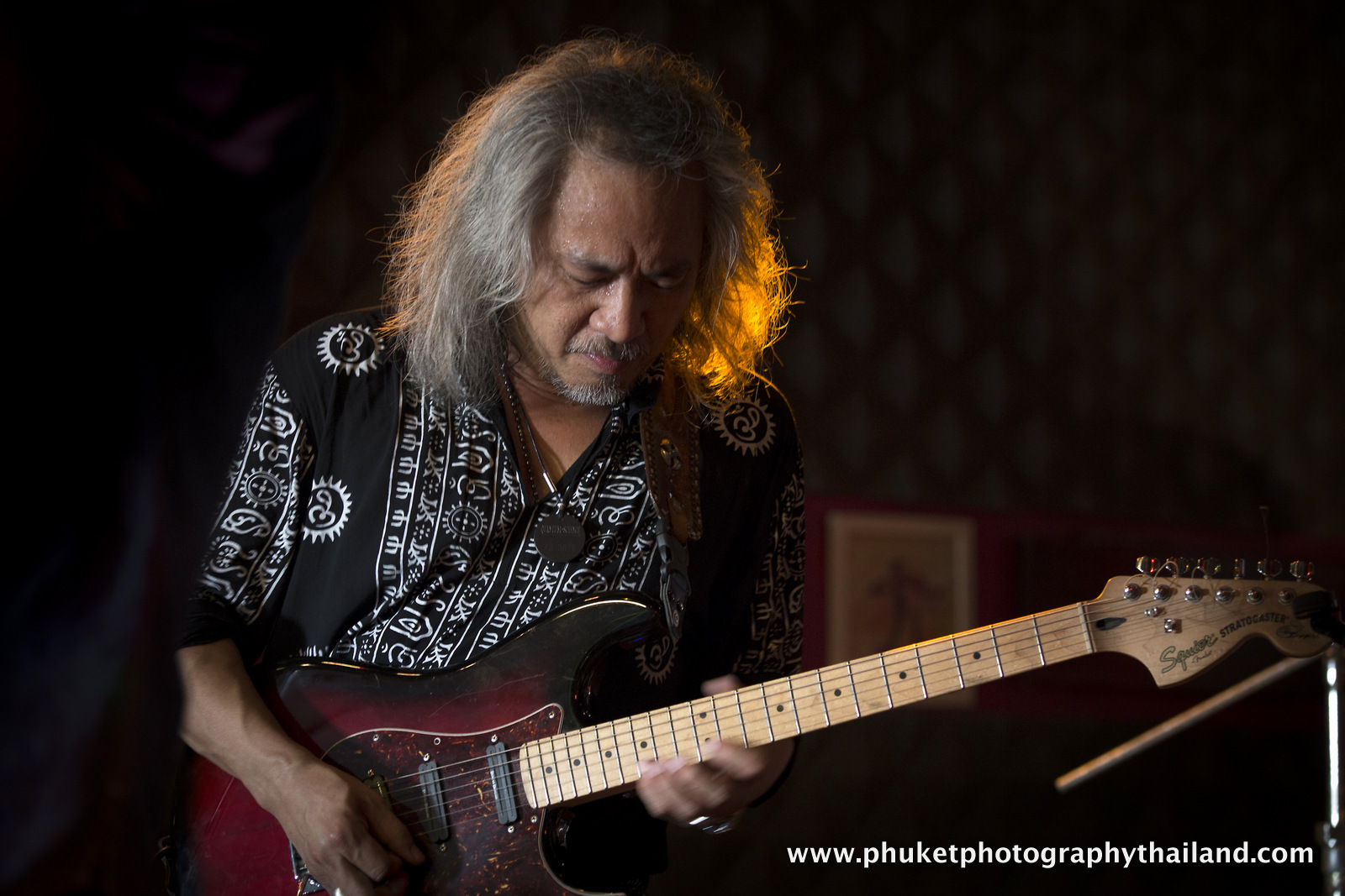A musician with long, curly hair performs passionately on an electric guitar in a dimly lit venue.