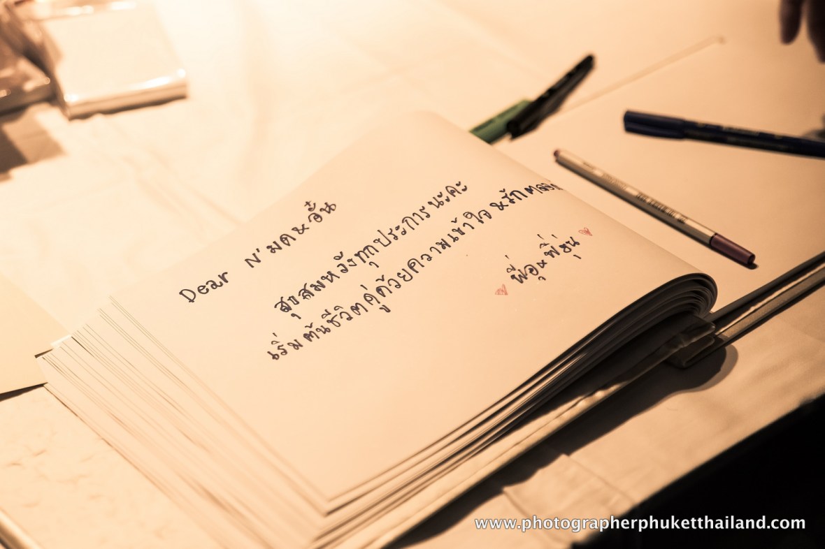 A close-up of a wedding guestbook with handwritten messages in both English and Thai, accompanied by pens on a table.