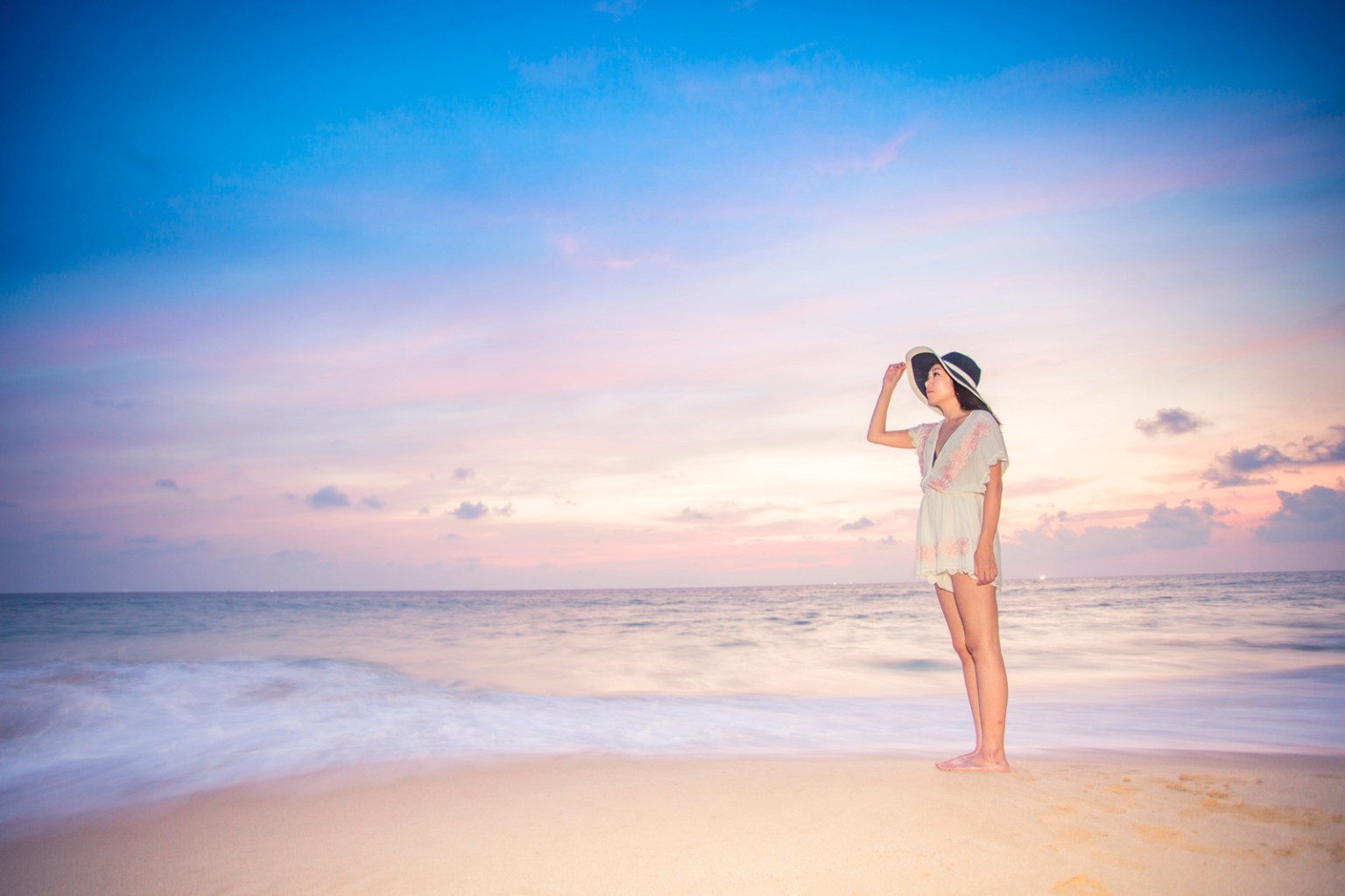 A person standing on the beach at sunset, wearing a stylish hat and a light-colored outfit, gazing out towards the ocean with a pink and blue sky in the background.