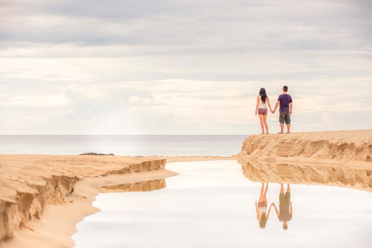 A couple standing hand in hand on a sandy beach, facing the ocean with a calm sky. Their reflections are visible in a shallow pool of water beside them.