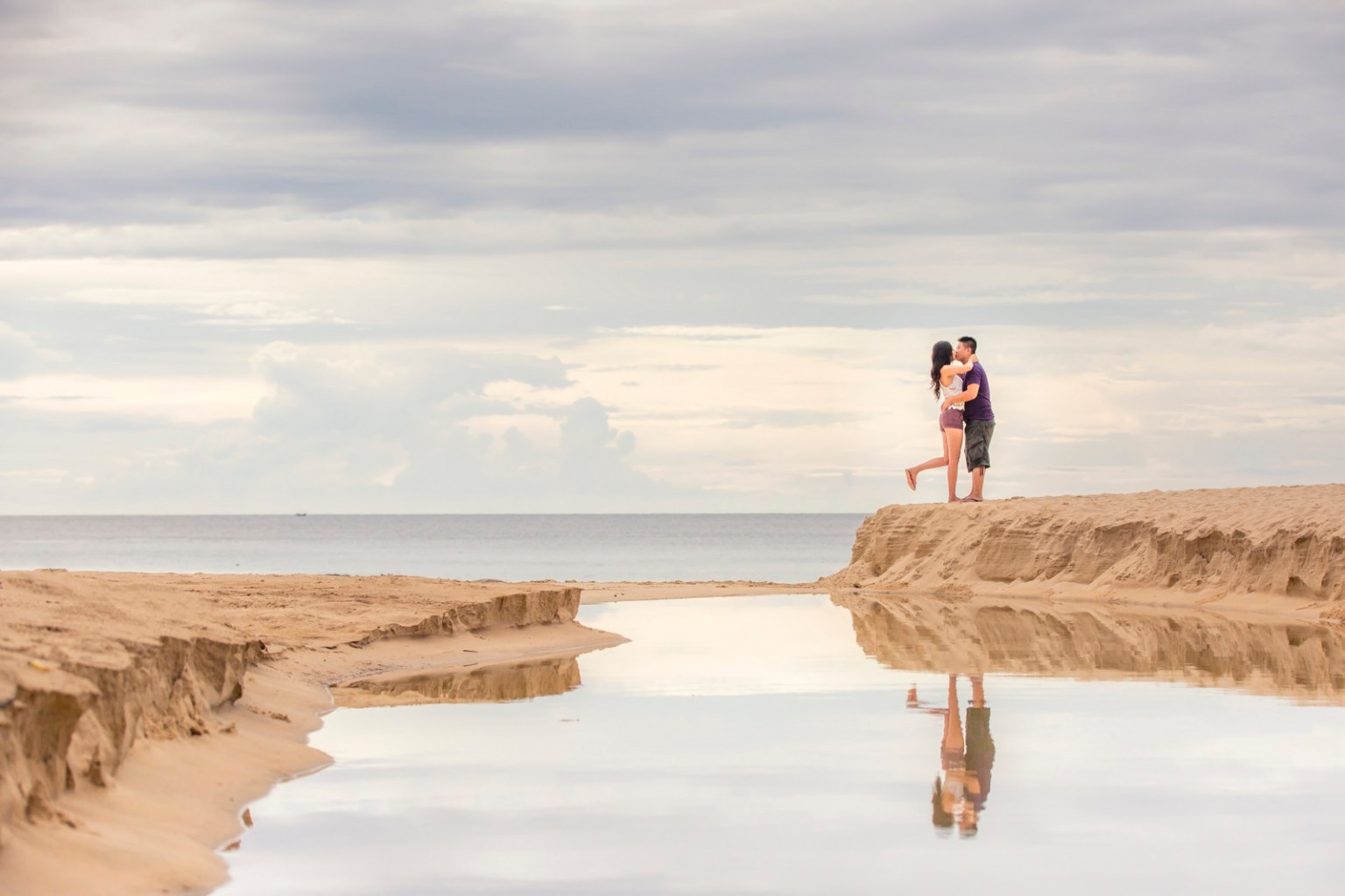 A couple embracing on a sandy beach near the water, reflected in a shallow pool of water with a cloudy sky above at surin beach phuket.