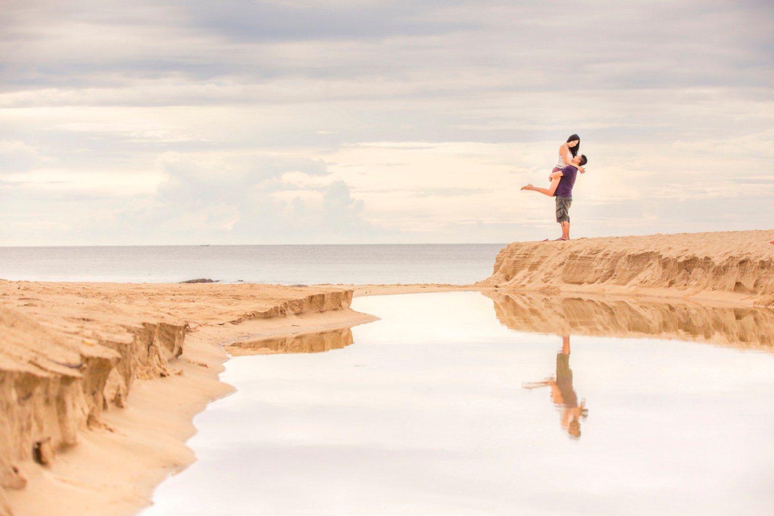 A couple embracing on a sandy beach with a calm sea in the background, reflecting the sky and surrounding scenery.