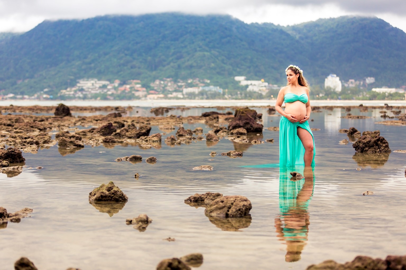 A pregnant woman in a turquoise outfit stands in shallow water surrounded by rocks, with a mountainous landscape in the background.