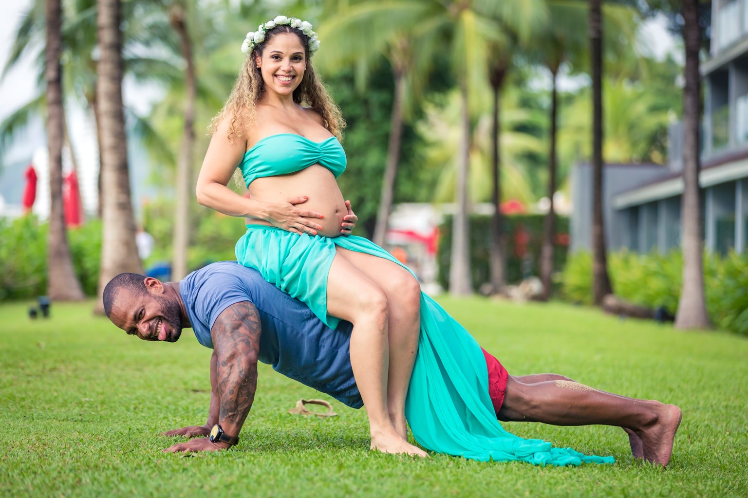 A pregnant woman in a turquoise dress sits on the back of a man doing push-ups in a lush green outdoor setting with palm trees.