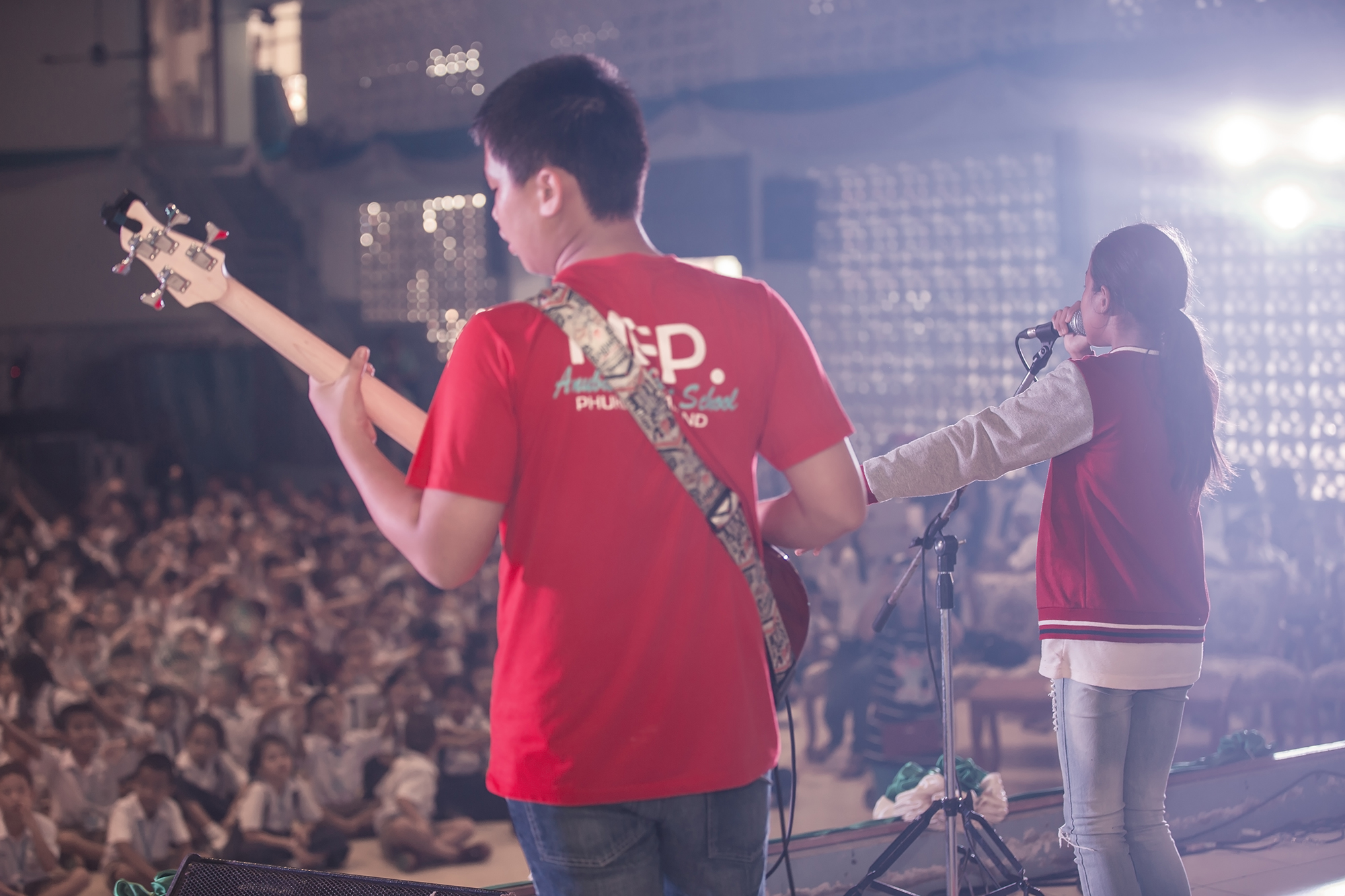 A live music performance featuring a guitarist in a red shirt and a singer in a red and gray jacket, with an audience in the background.