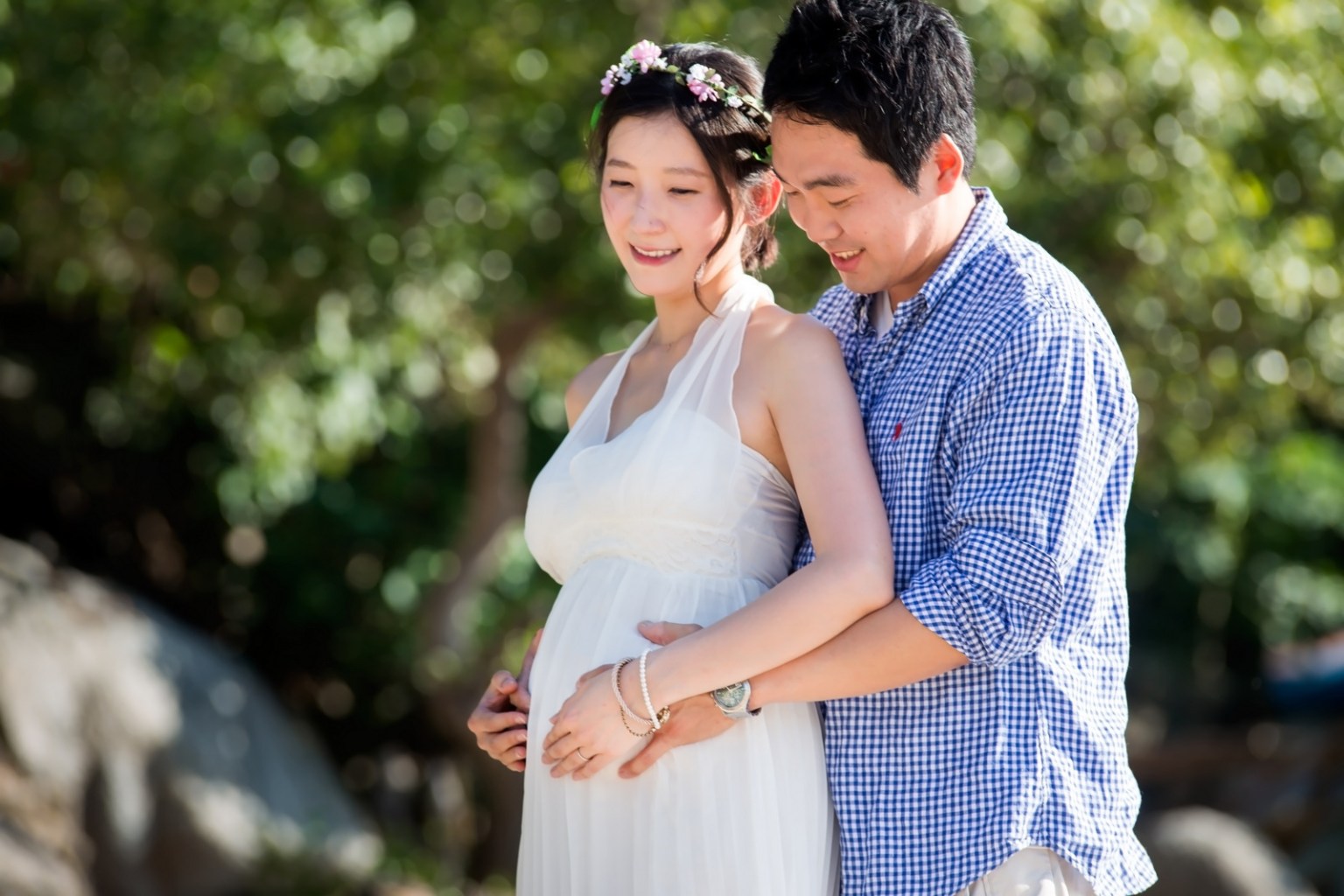 A couple poses together outdoors, with the woman wearing a white dress and floral crown, cradling her baby bump while the man stands behind her, smiling.