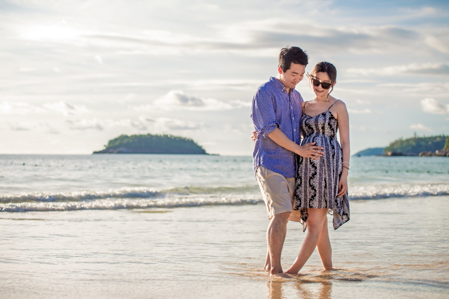 A couple standing together on a sandy beach, with the woman cradling her baby bump and wearing a sundress, while the man in a checkered shirt stands beside her, both smiling at each other. The ocean and an island are visible in the background under a bright sky.