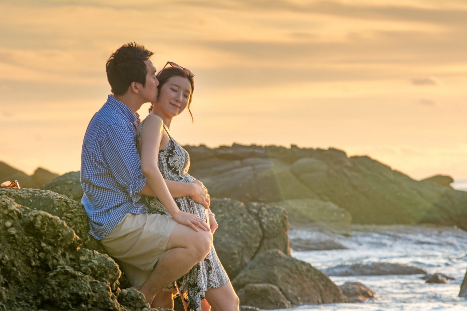 A couple enjoying a romantic moment on the rocks by the beach during sunset, with gentle waves in the background.