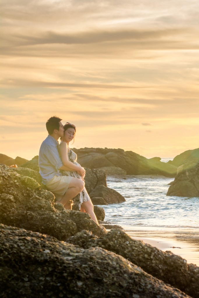 A couple sitting on rocks by the beach during sunset, with the ocean and clouds in the background.