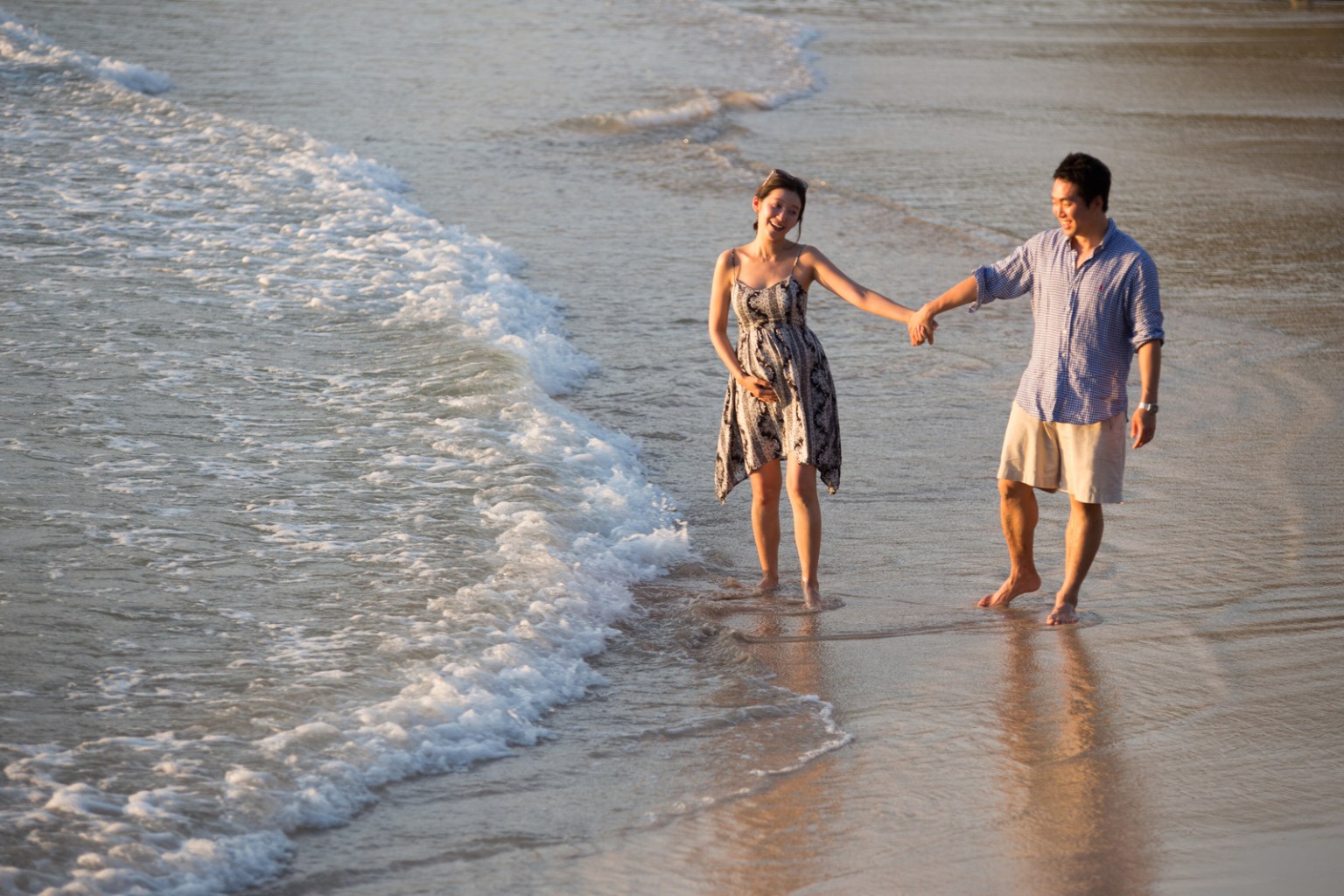 A couple walking hand in hand along the beach at sunset, with gentle waves lapping at their feet.
