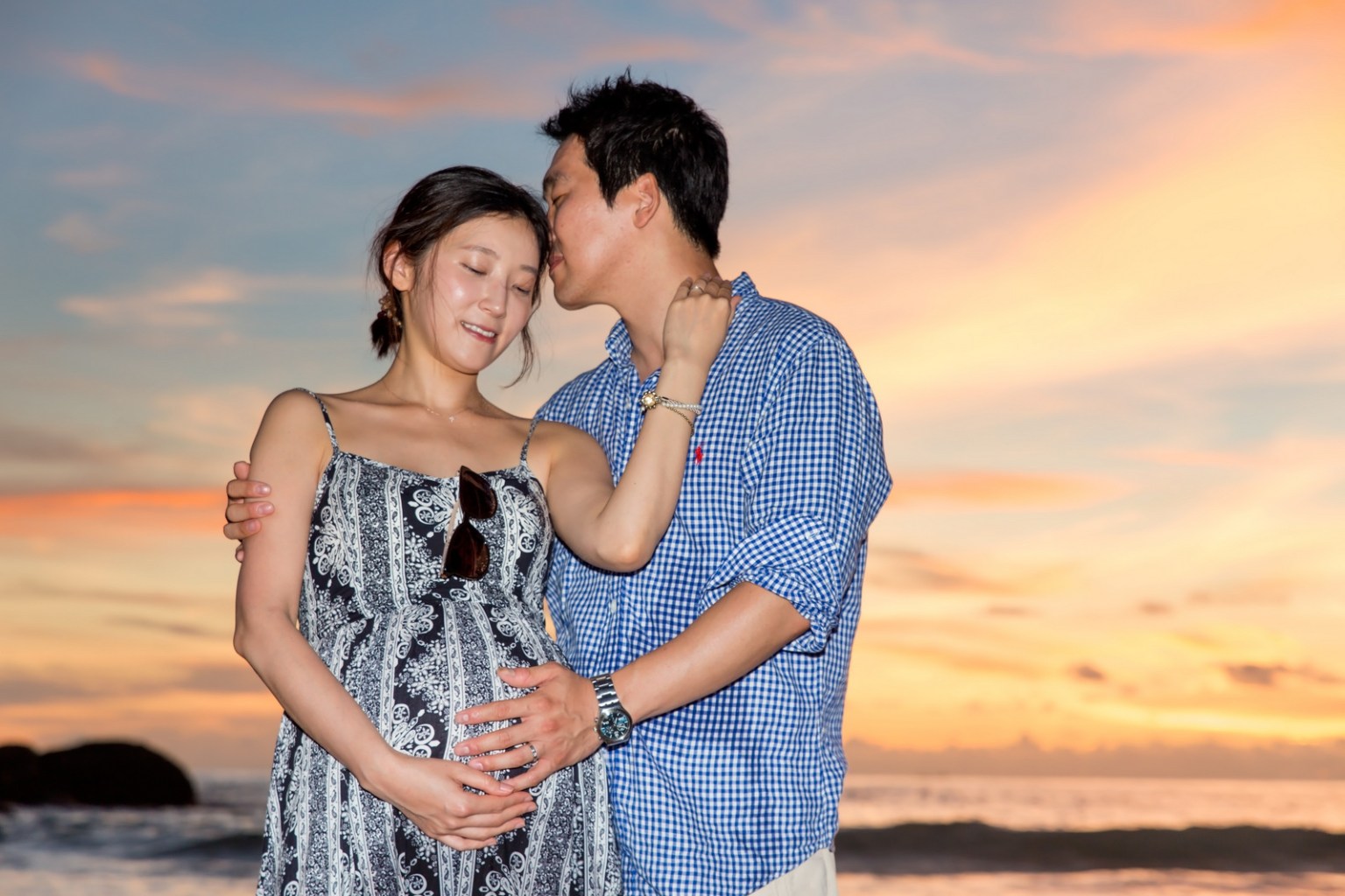 A couple embracing at the beach during sunset, with the woman showing her baby bump.
