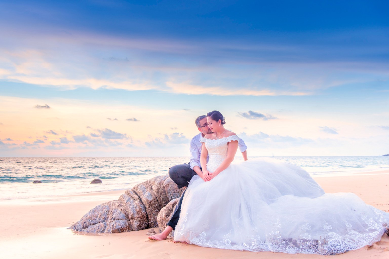 A couple embraces on the beach during sunset, with the woman wearing a wedding dress and the man in a light shirt, sitting on a rock by the sea.