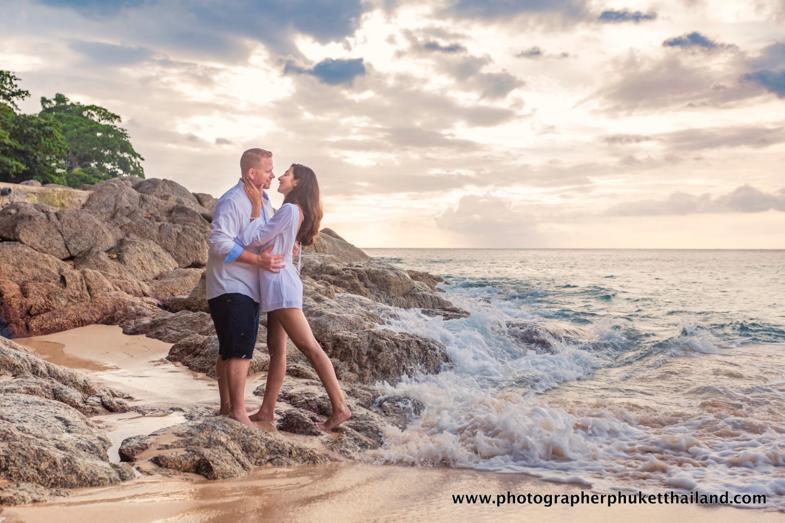A couple embracing on rocky beach during sunset, with waves crashing around them.