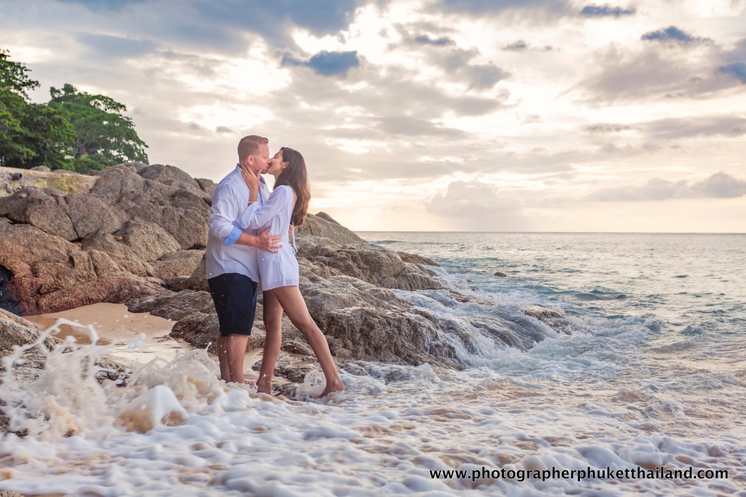 A couple kissing on a rocky beach with waves crashing around them and a cloudy sky in the background.