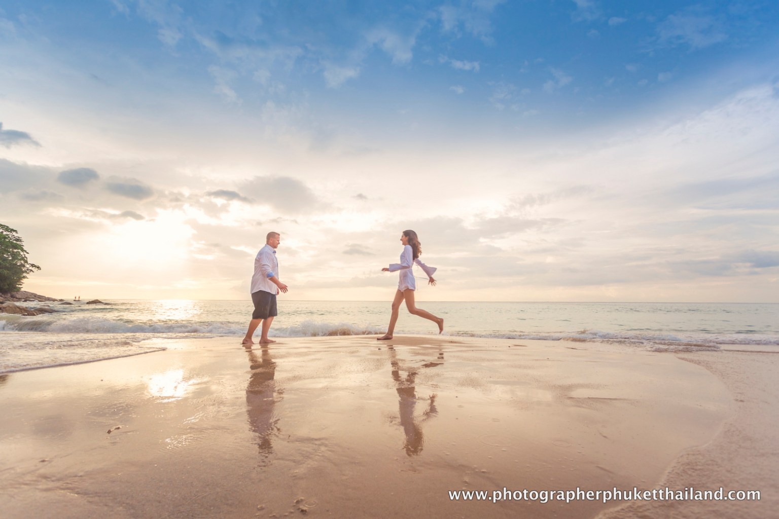 A couple is playfully running towards each other on a beach at sunset, with the ocean waves in the background and a colorful sky.