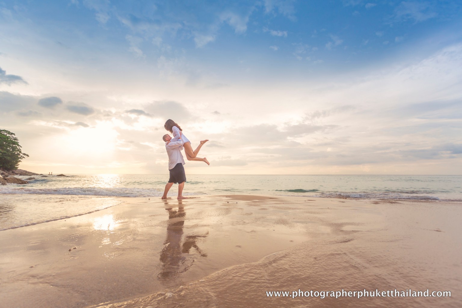 A couple at surin beach joyfully enjoying a moment on the beach, with the sun setting in the background and the ocean gently lapping at their feet.