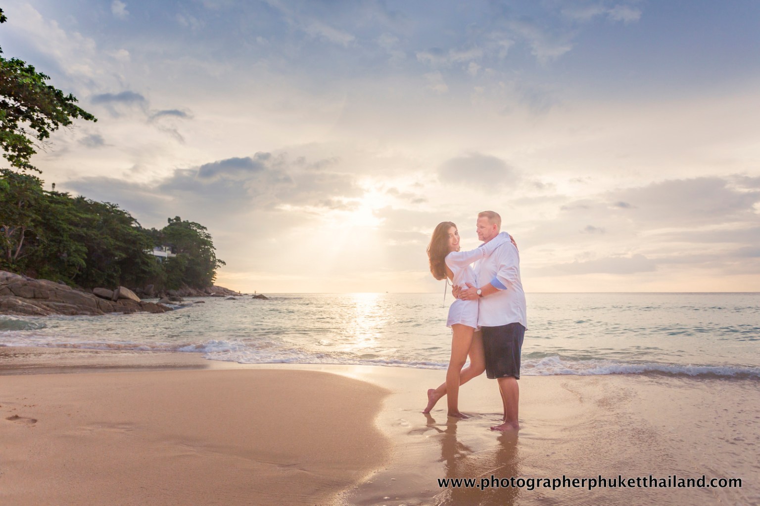 A couple embracing on a beach during sunset, with the ocean in the background and gentle waves lapping at the shore.