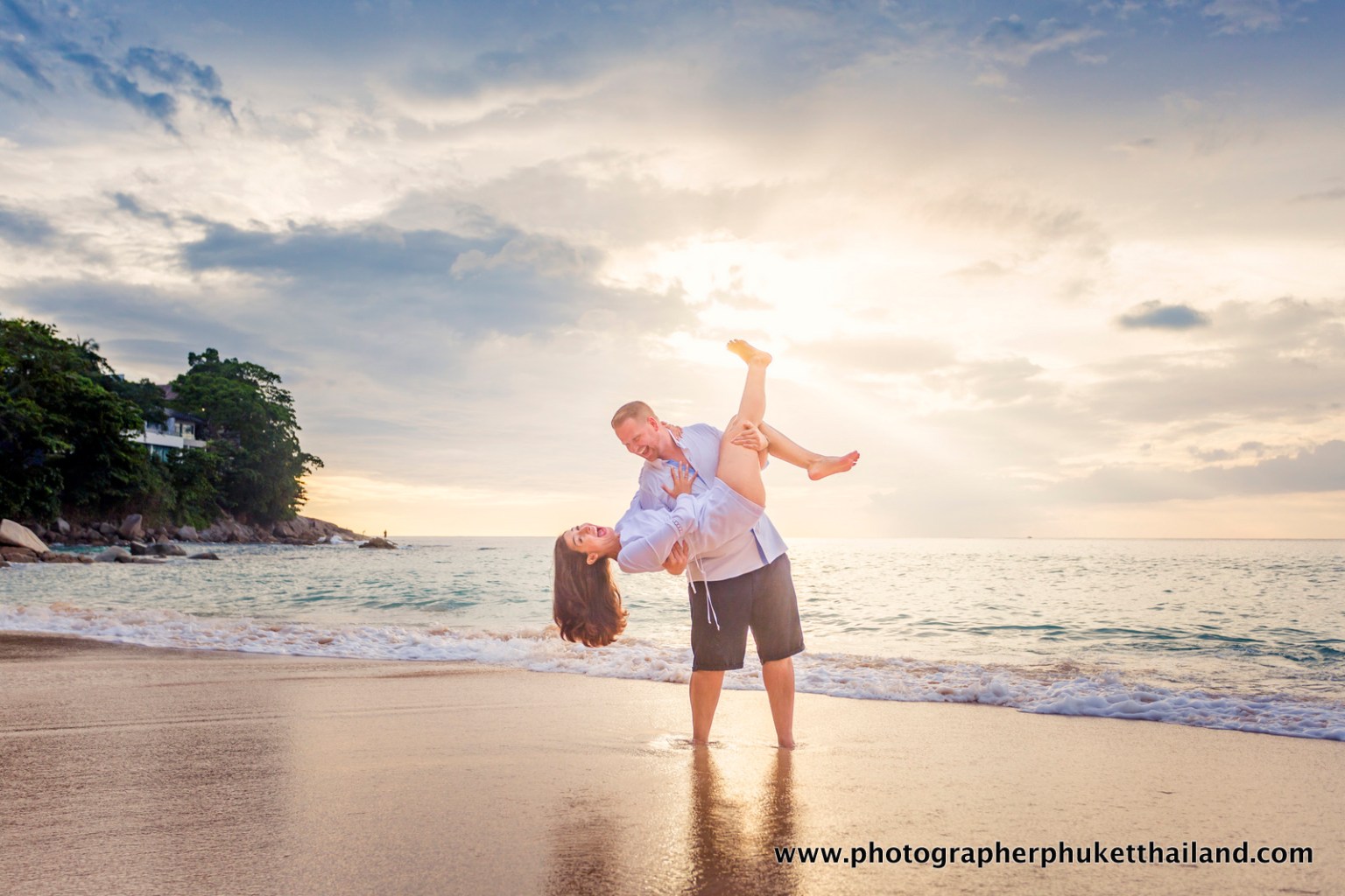 A couple playfully embracing on a beach at sunset, with the woman being lifted by the man. The sky is partly cloudy and the ocean waves can be seen in the background.