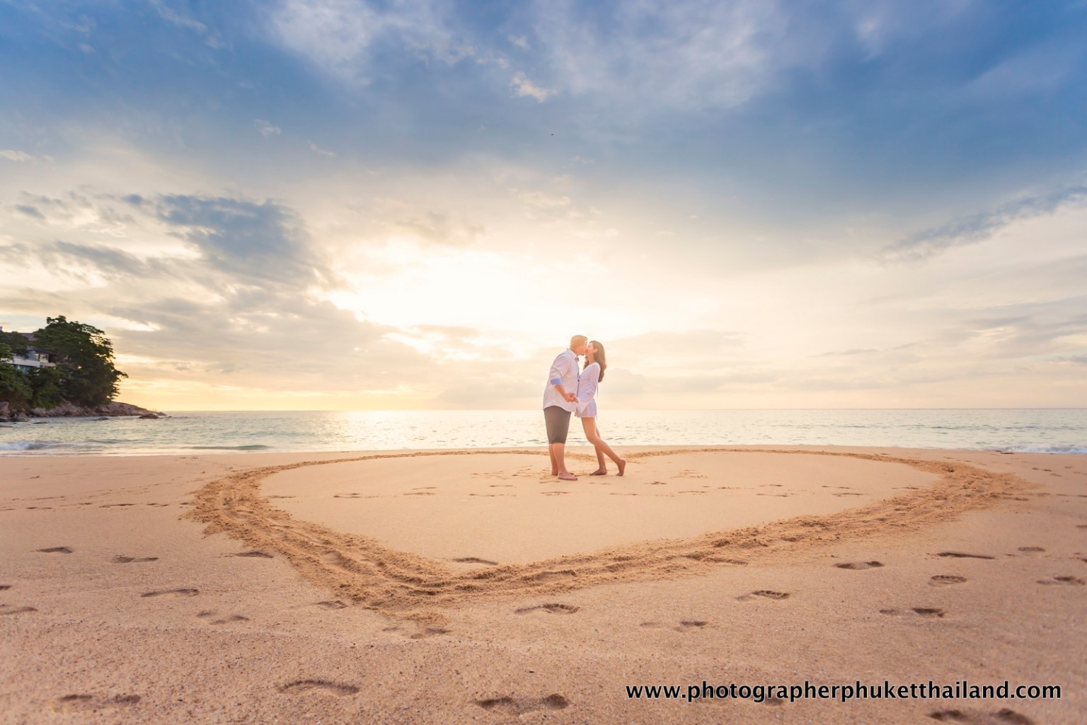 A couple kissing in a heart-shaped area on the beach during sunset, with a beautiful sky and ocean in the background.
