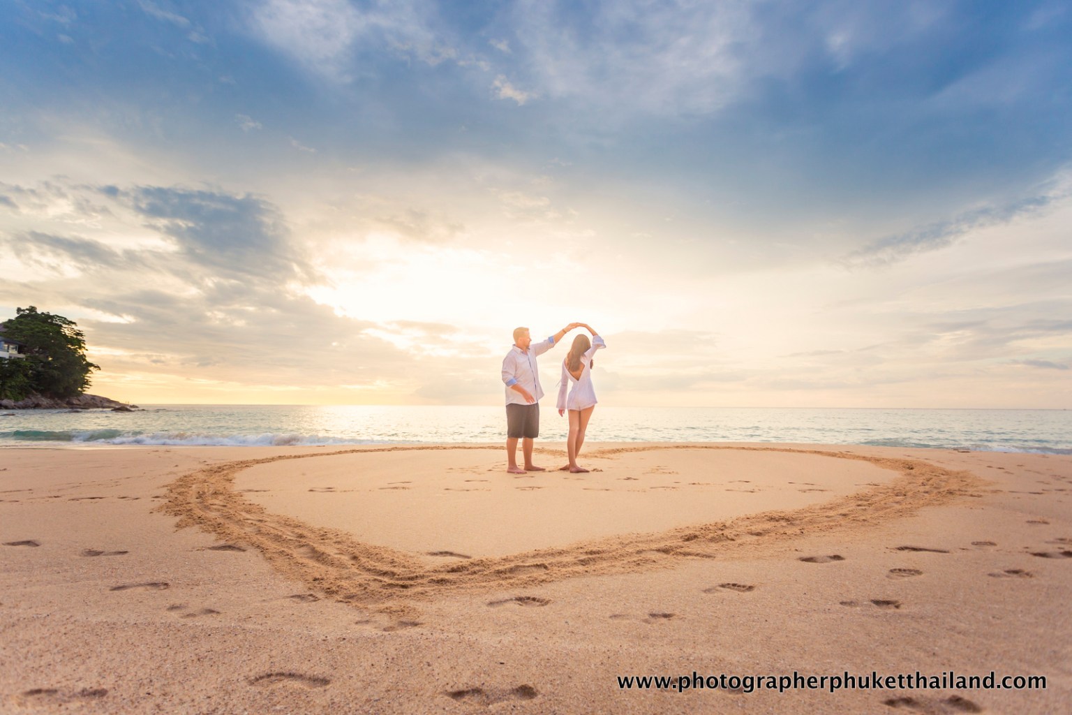 A couple dancing on the surin beach at sunset, with their feet in the sand and a heart shape drawn in the sand.
