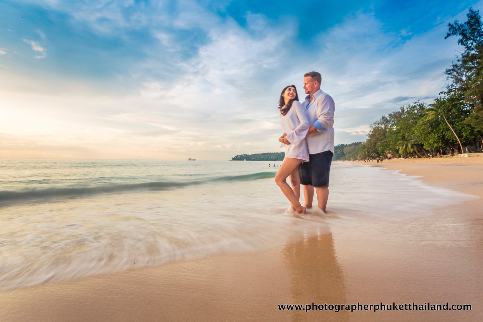 A couple standing together on a beach at sunset, with the waves gently lapping at their feet.