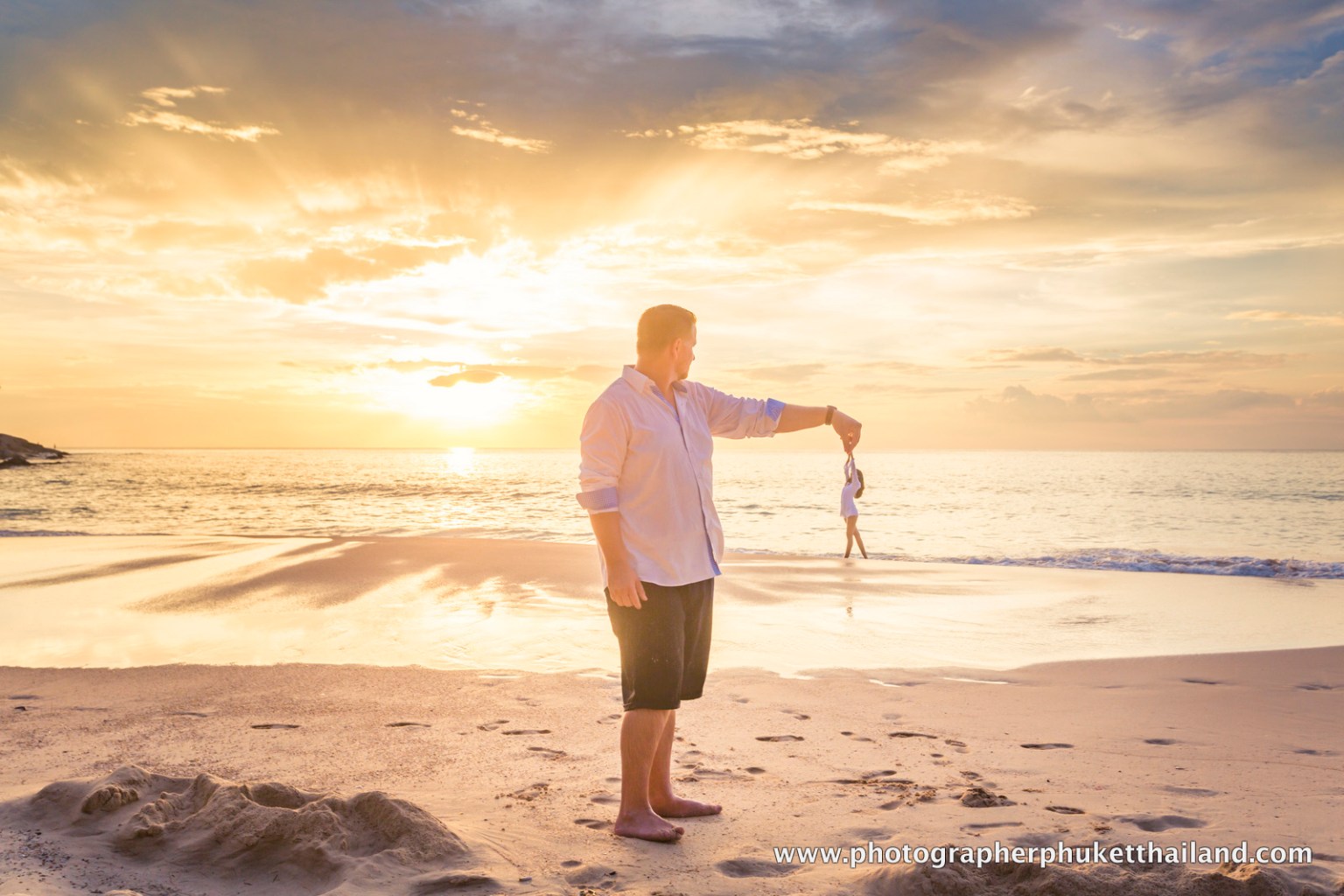A man in a white shirt holds a piece of fabric while standing on a beach during sunset, with another person walking in the background.