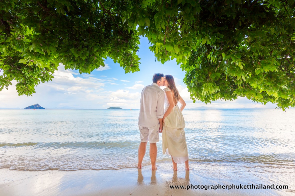A couple kisses on a beach under lush green foliage, with calm water and distant islands in the background.