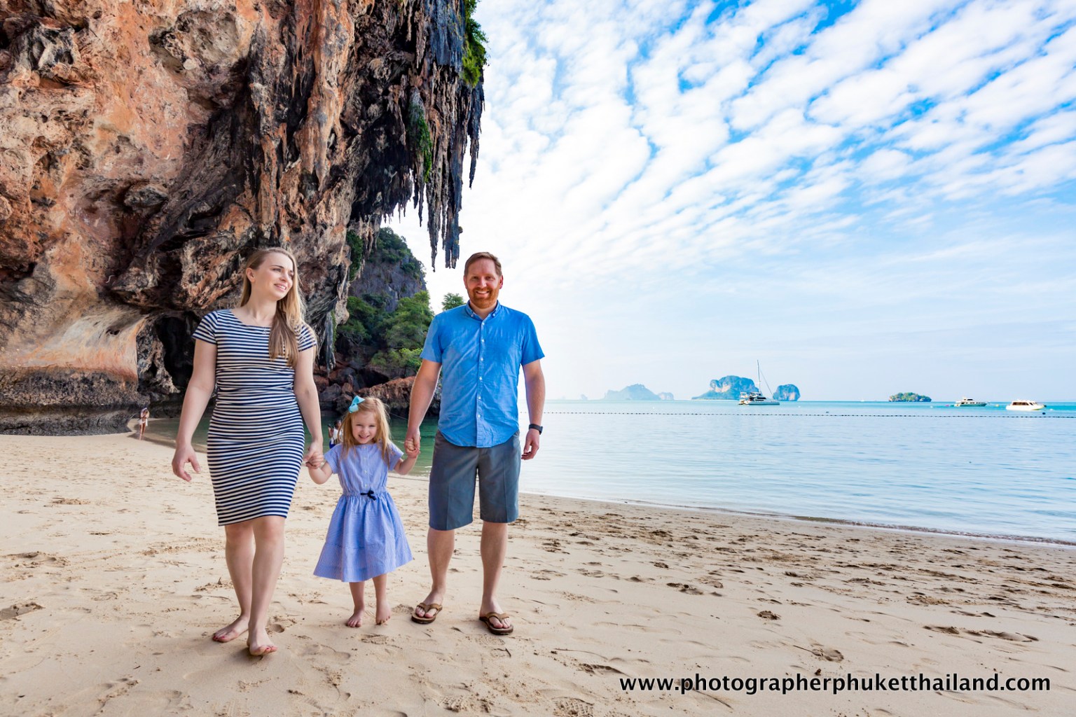 A family of three, including a woman, a man, and a girl, walking hand in hand on a sandy beach with large limestone cliffs in the background and a clear blue sky.