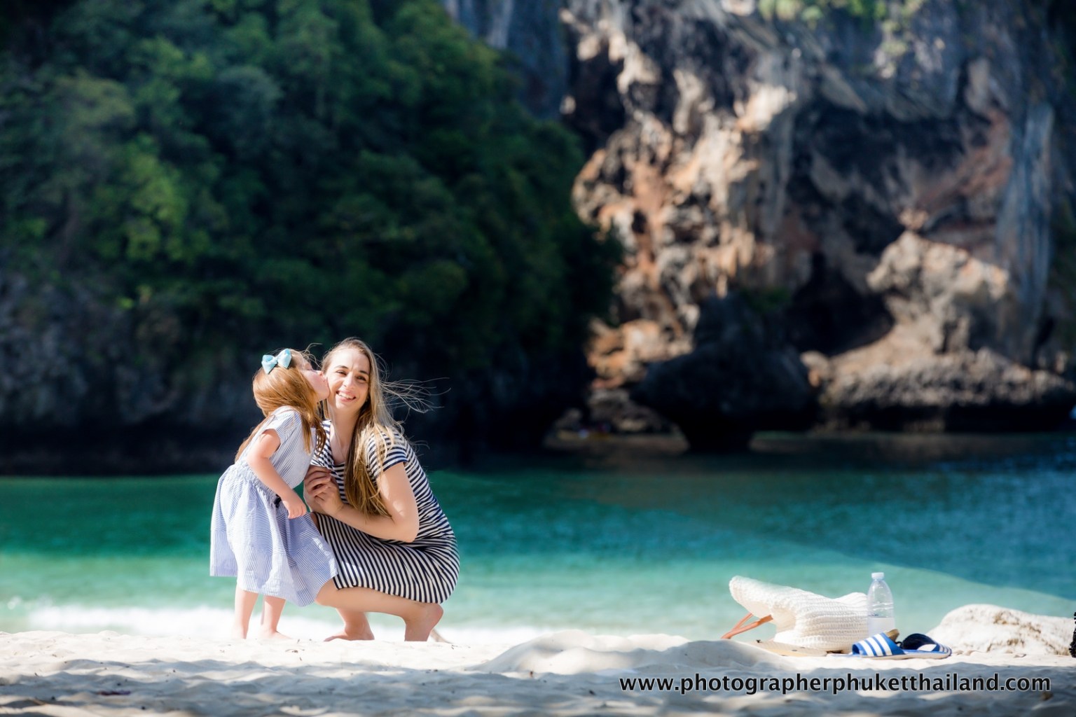 A woman and a young girl sharing a joyful moment on the beach in Krabi, Thailand, with limestone cliffs in the background.