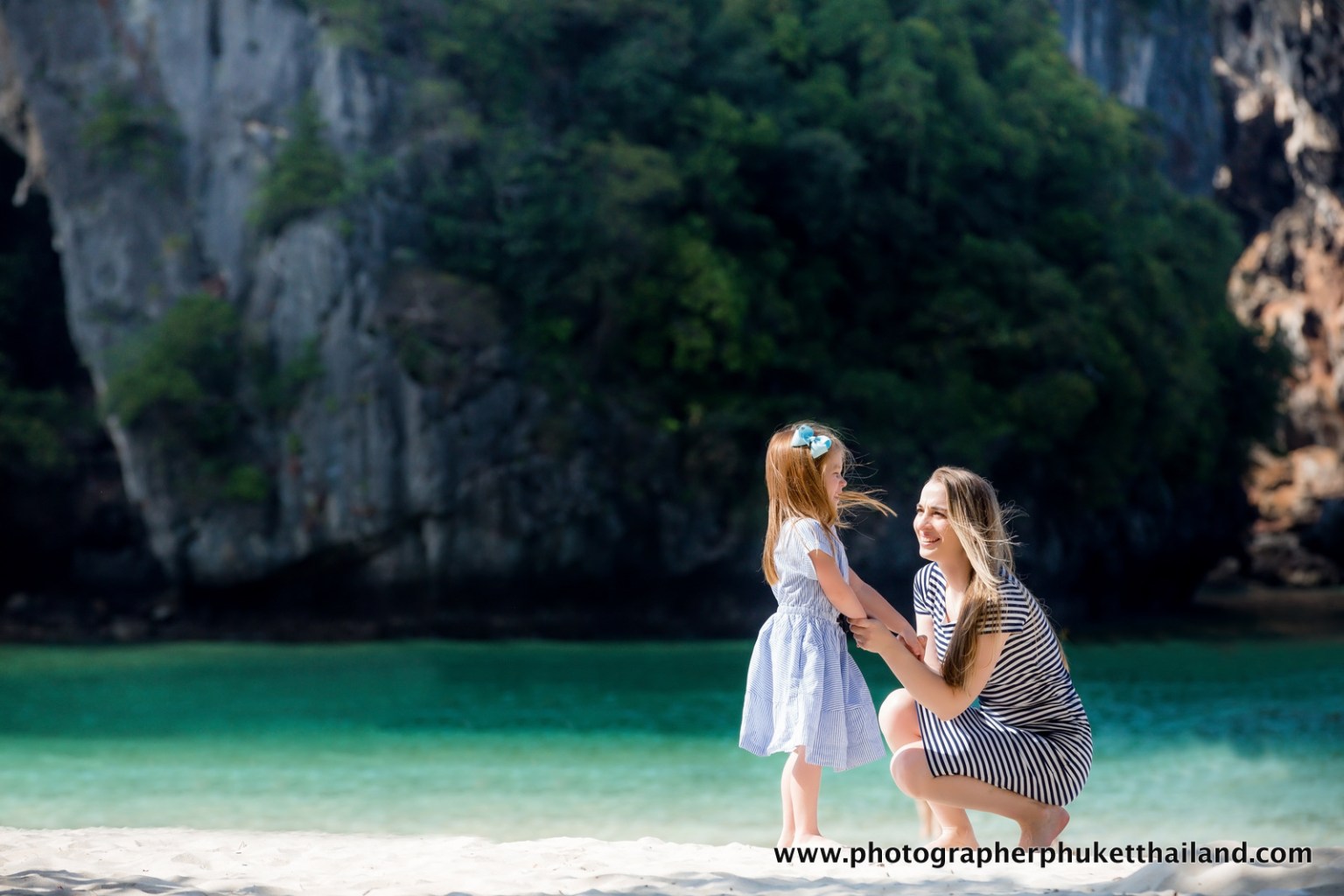 A woman and a young girl play on a sandy beach near crystal-clear waters, surrounded by towering limestone cliffs covered in greenery, with sunlight illuminating the scene.