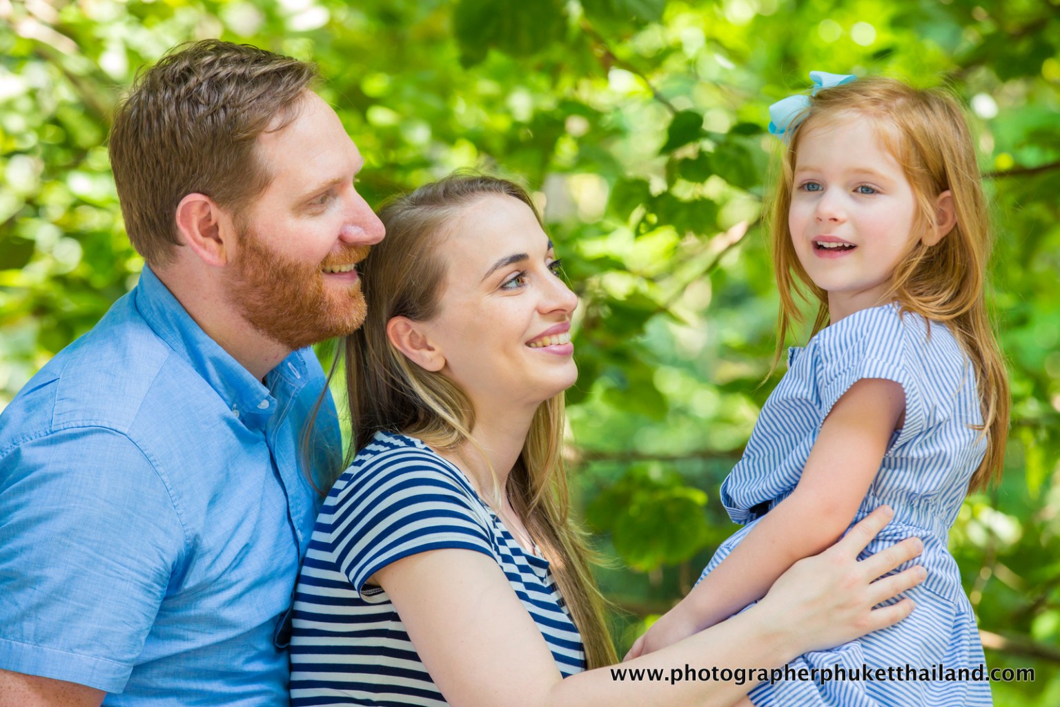 Happy family portrait with a couple and their young daughter, smiling together in a lush green setting.