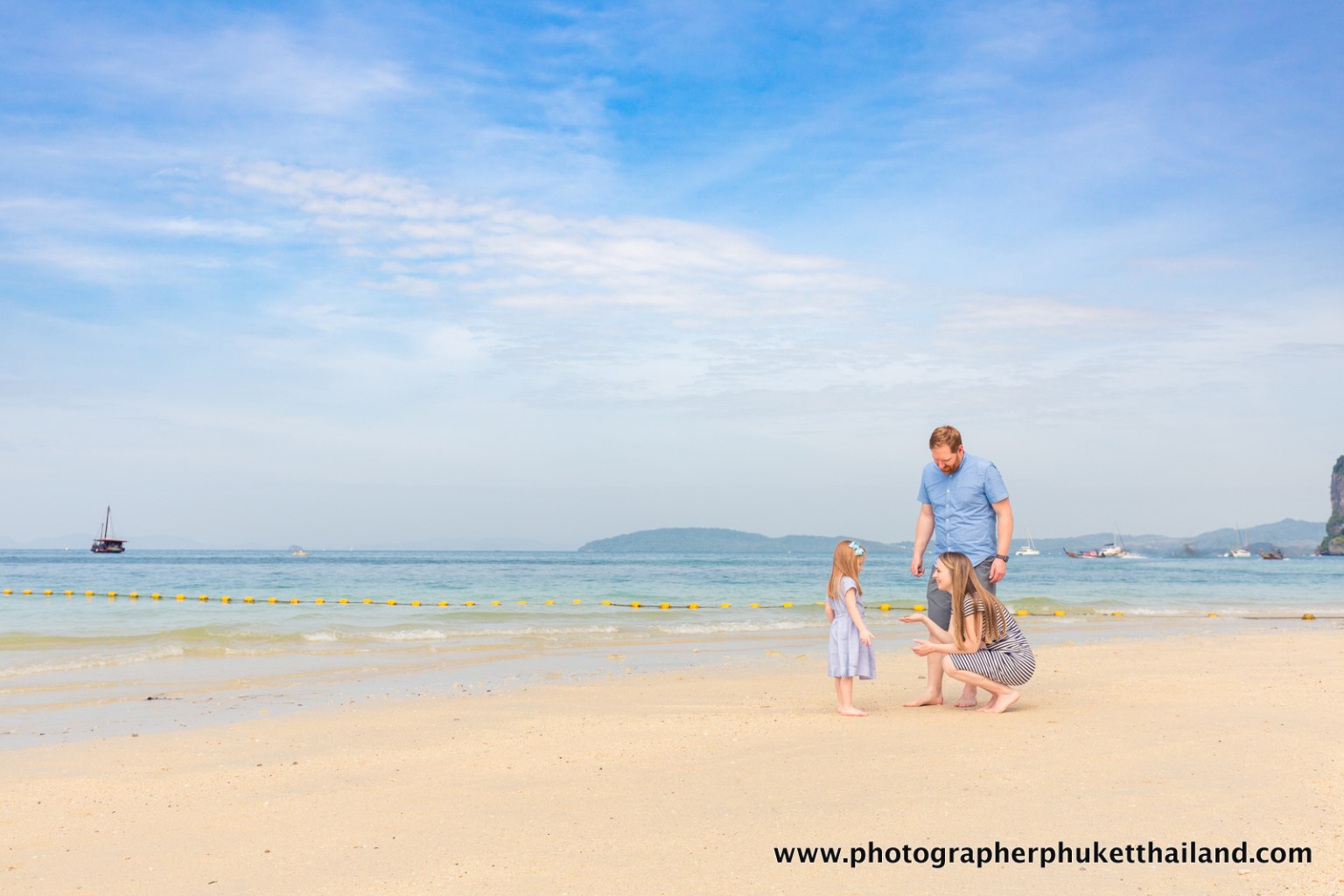 A family at the beach in Krabi, Thailand, with a couple interacting with their young daughter on the sandy shore and a boat in the background.
