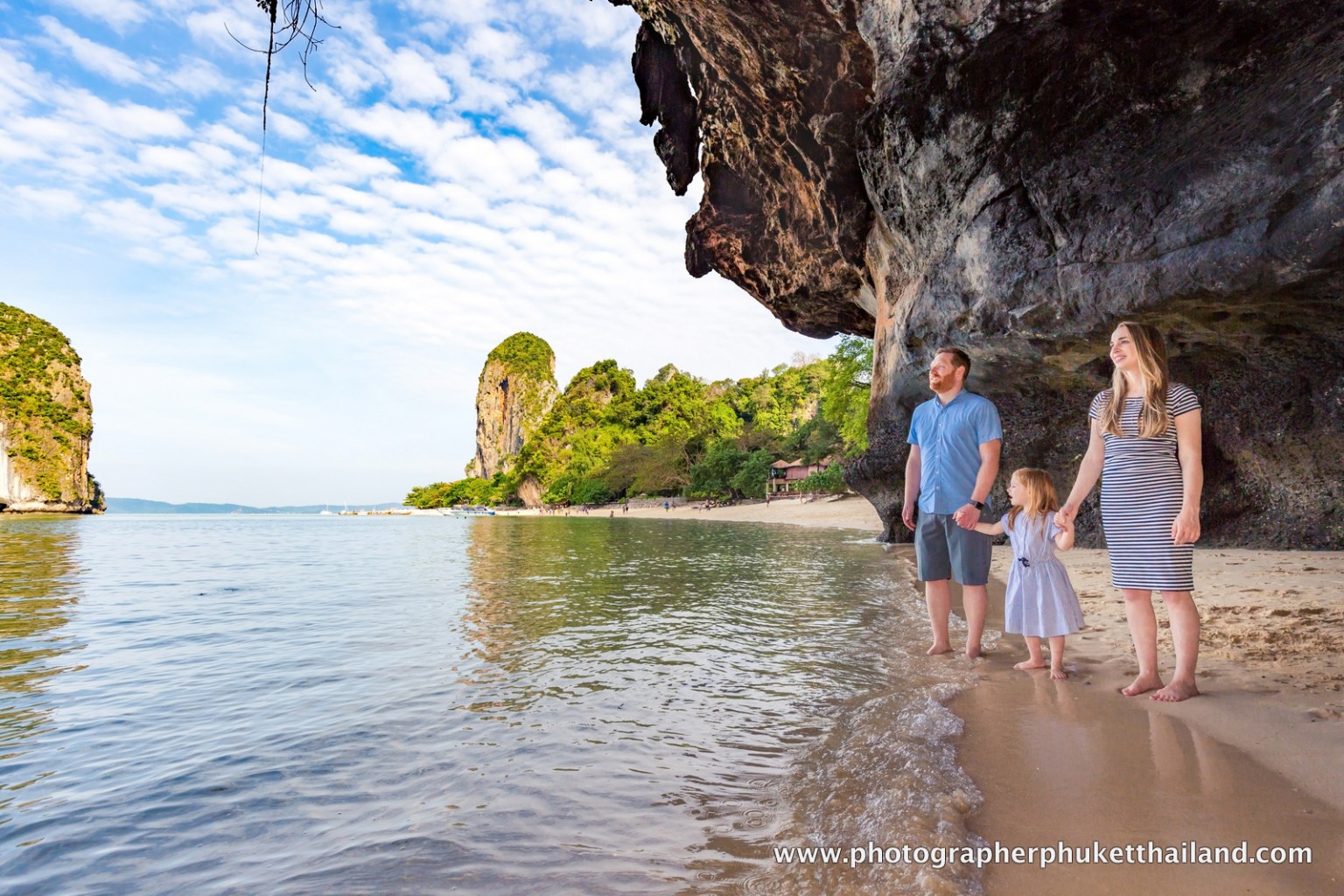 A couple and a child standing on a beach in Krabi, Thailand, with limestone cliffs and a clear sky in the background.
