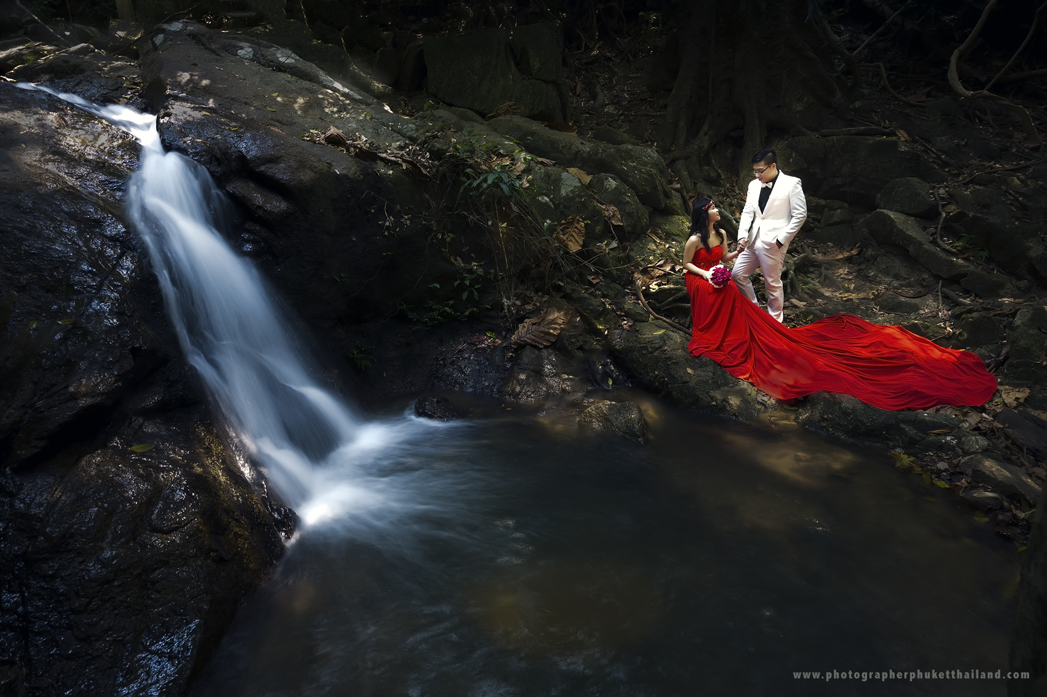 A couple poses near a waterfall, featuring the woman in a flowing red gown and the man in a white suit, set in a natural, leafy environment.