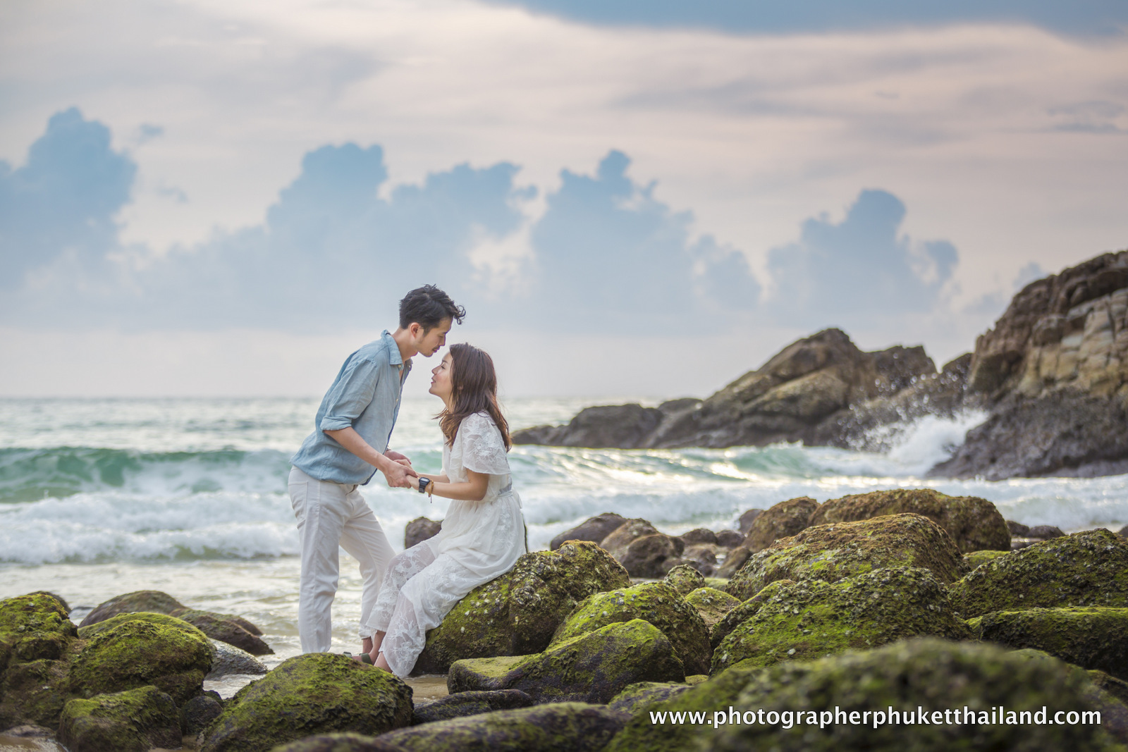A couple sharing a romantic moment on a rocky beach, with waves crashing in the background and a cloudy sky.