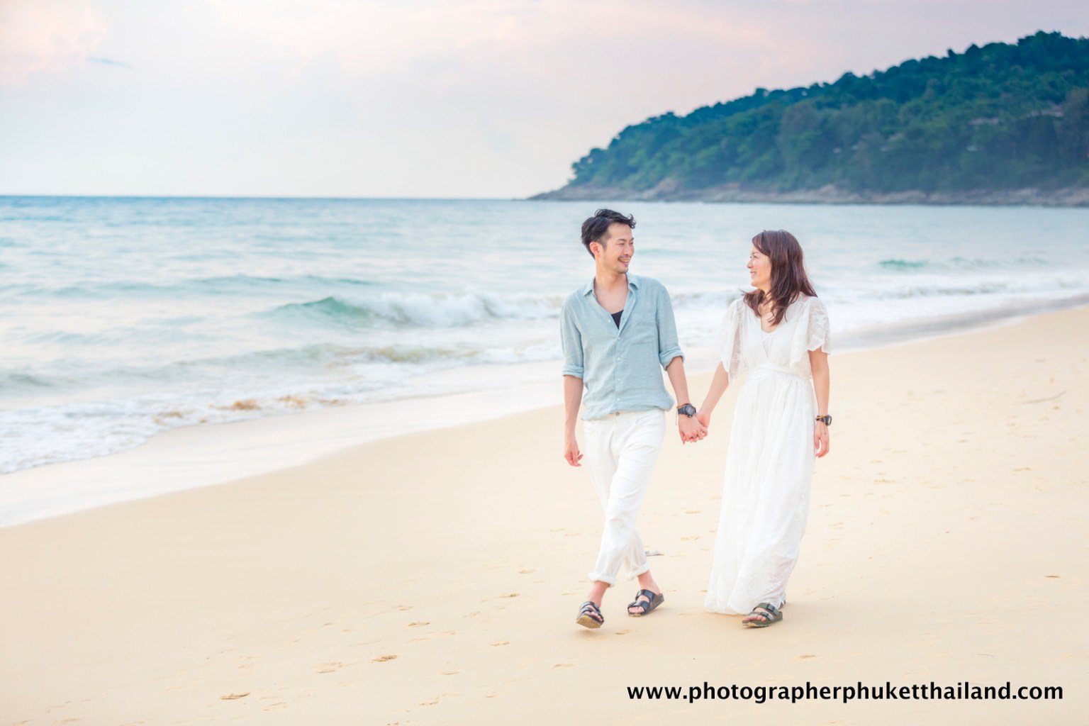 A couple walking hand-in-hand along the beach at sunset, with gentle waves lapping at the shore.