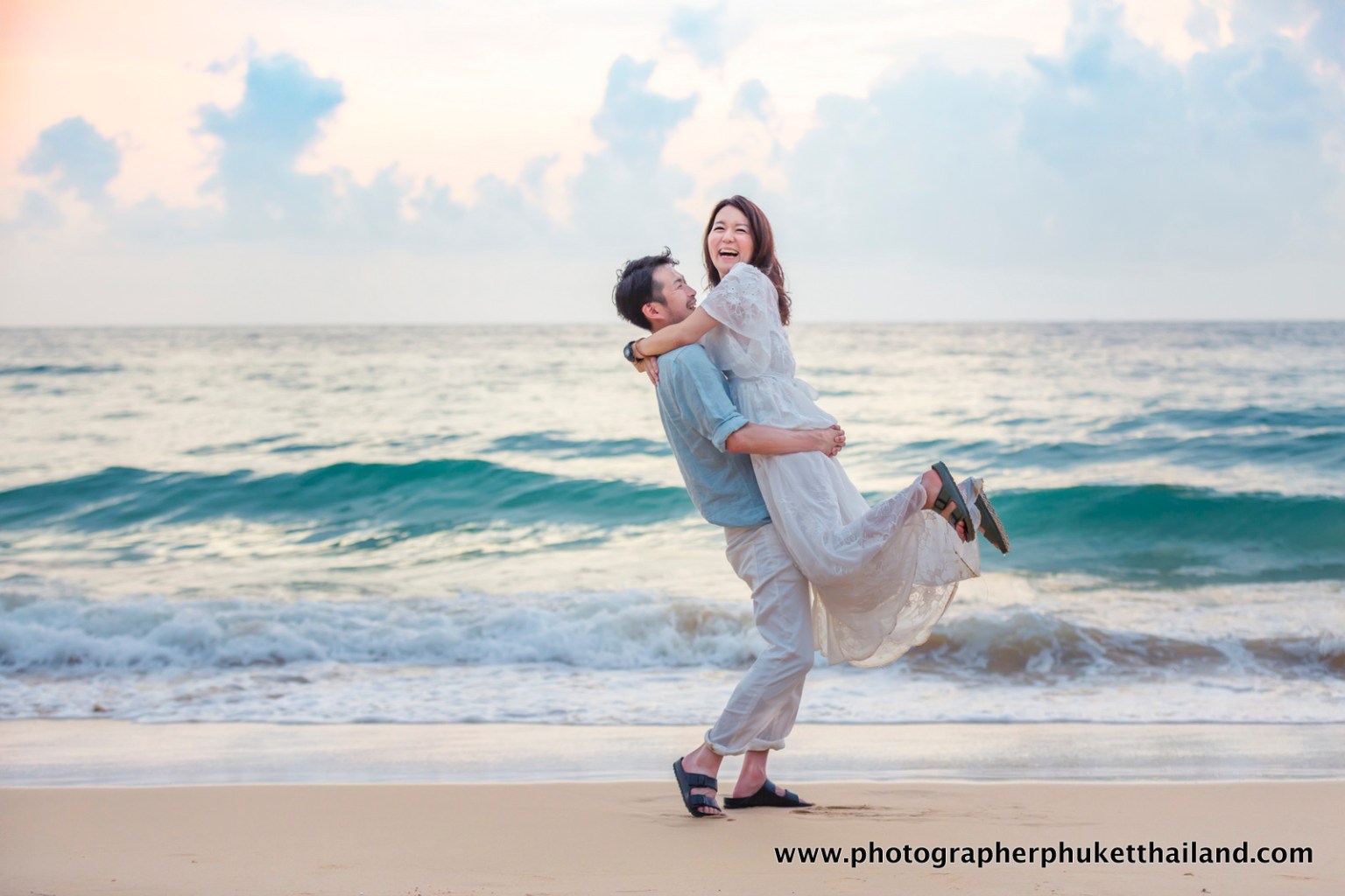 A joyful couple celebrating on the beach, with the man lifting the woman as they both smile. The sea waves crash in the background under a pastel sky.
