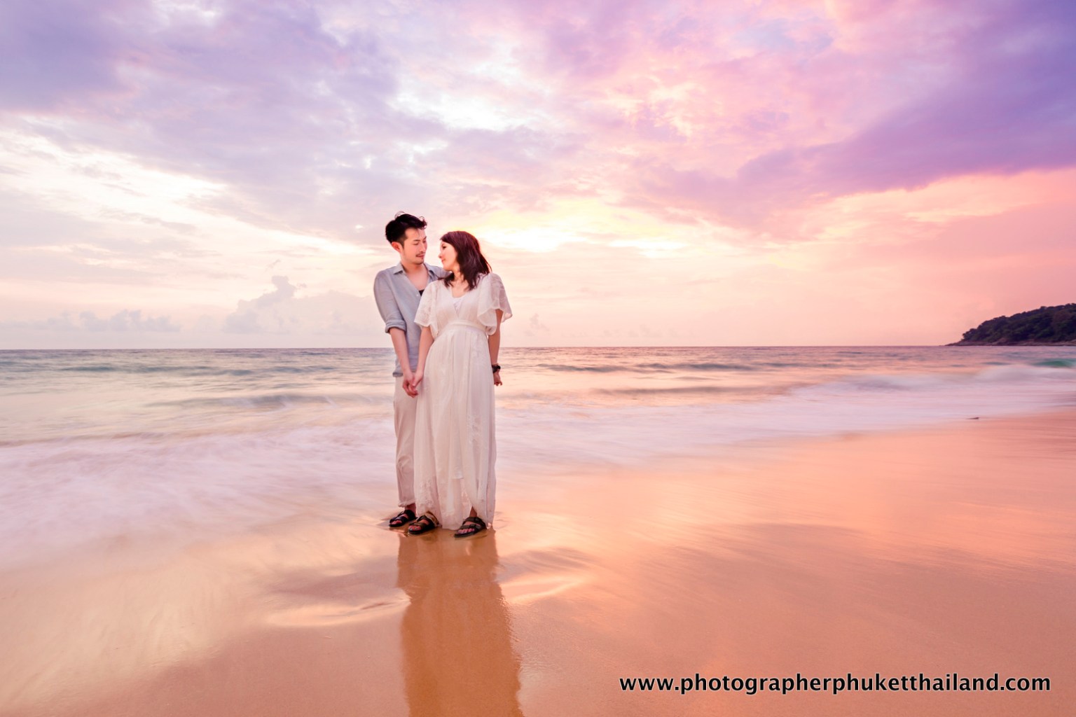 A couple stands on the beach holding hands at sunset, with colorful clouds reflecting on the water.