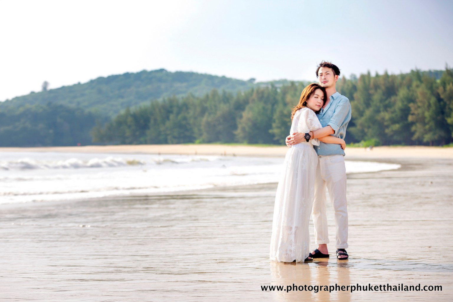 A couple embraces on the beach, with lush green hills and a calm sea in the background.