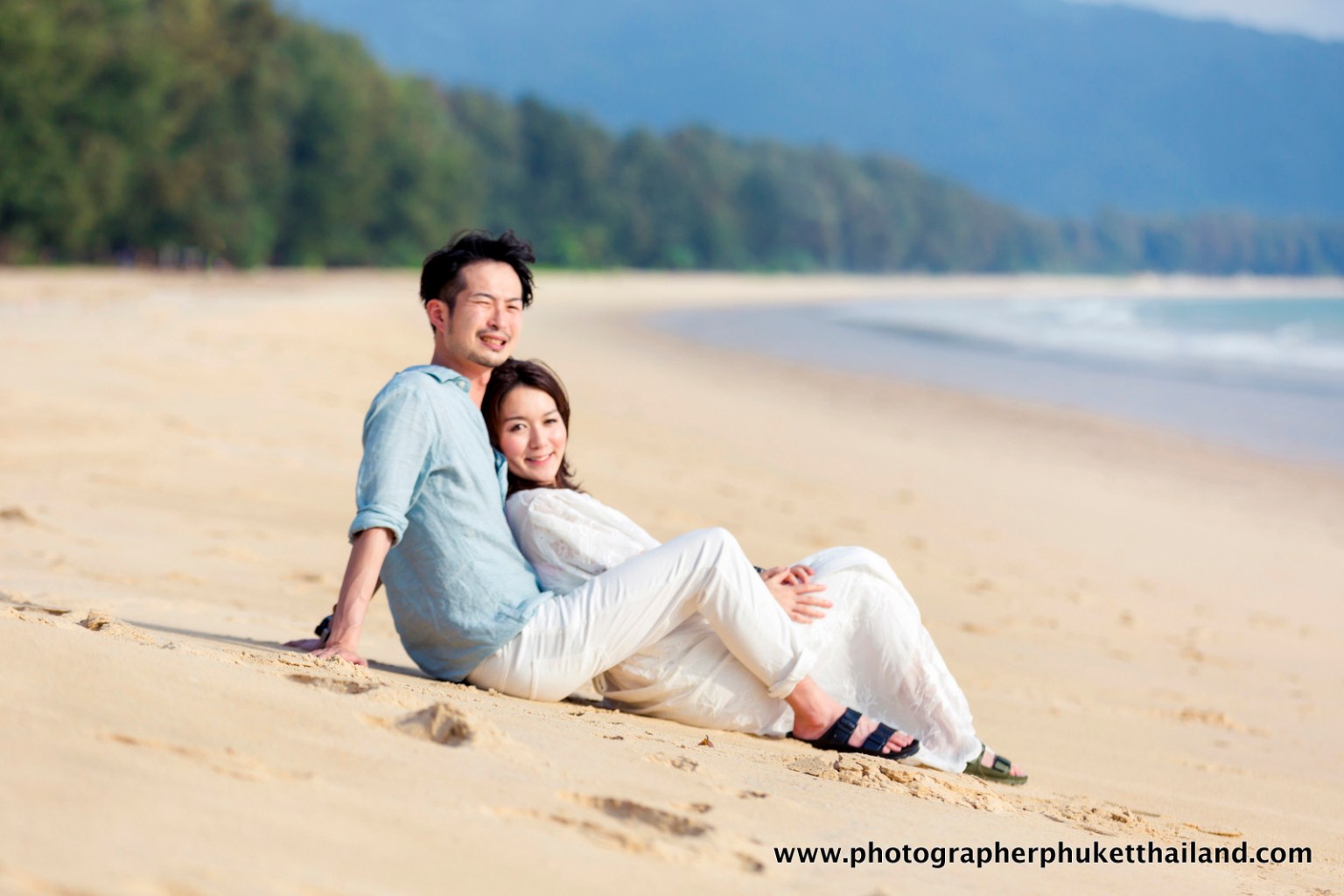 A couple sitting on a beach during sunset, smiling and enjoying each other's company, with the ocean in the background.
