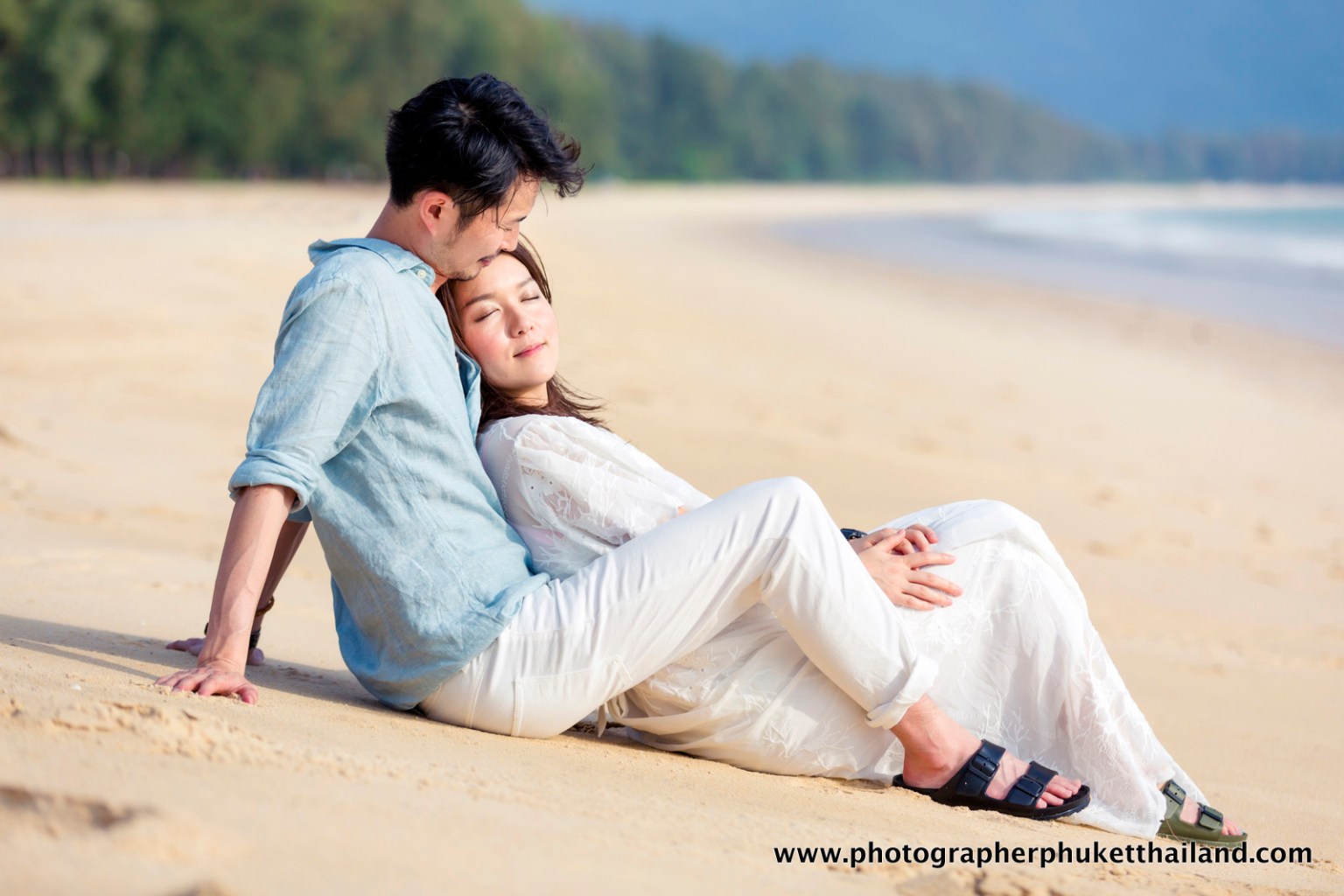 A couple sitting on the beach, with the woman resting her head on the man's shoulder, both gazing away with a serene expression. The setting features soft, sandy shores and a lush backdrop.