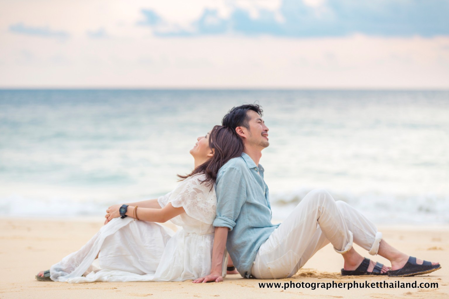 A romantic couple sitting back to back on a beach, smiling and enjoying the serene ocean view in the background.