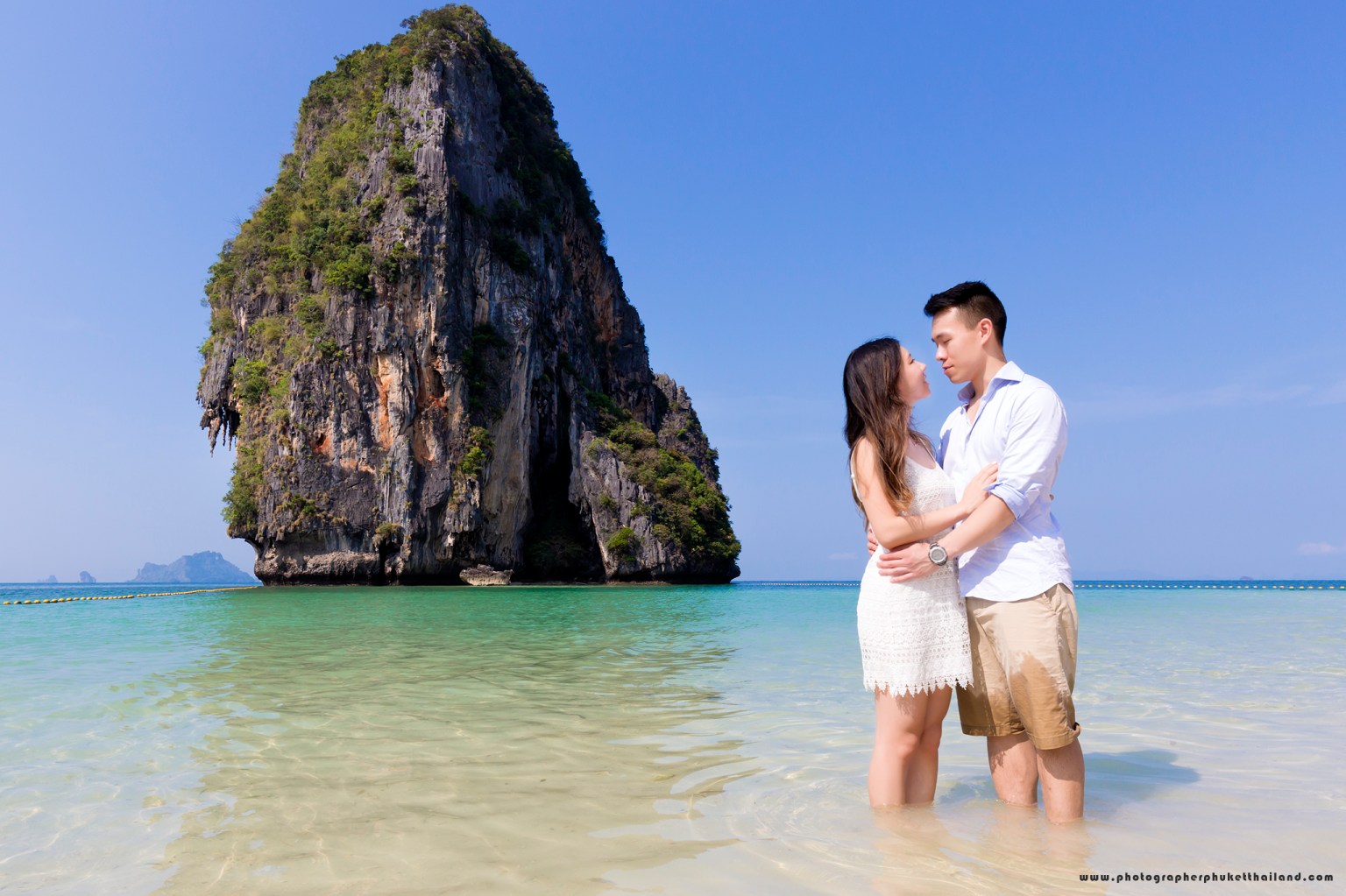 A couple embracing in shallow water on a sunny beach, with a large rock formation in the background.