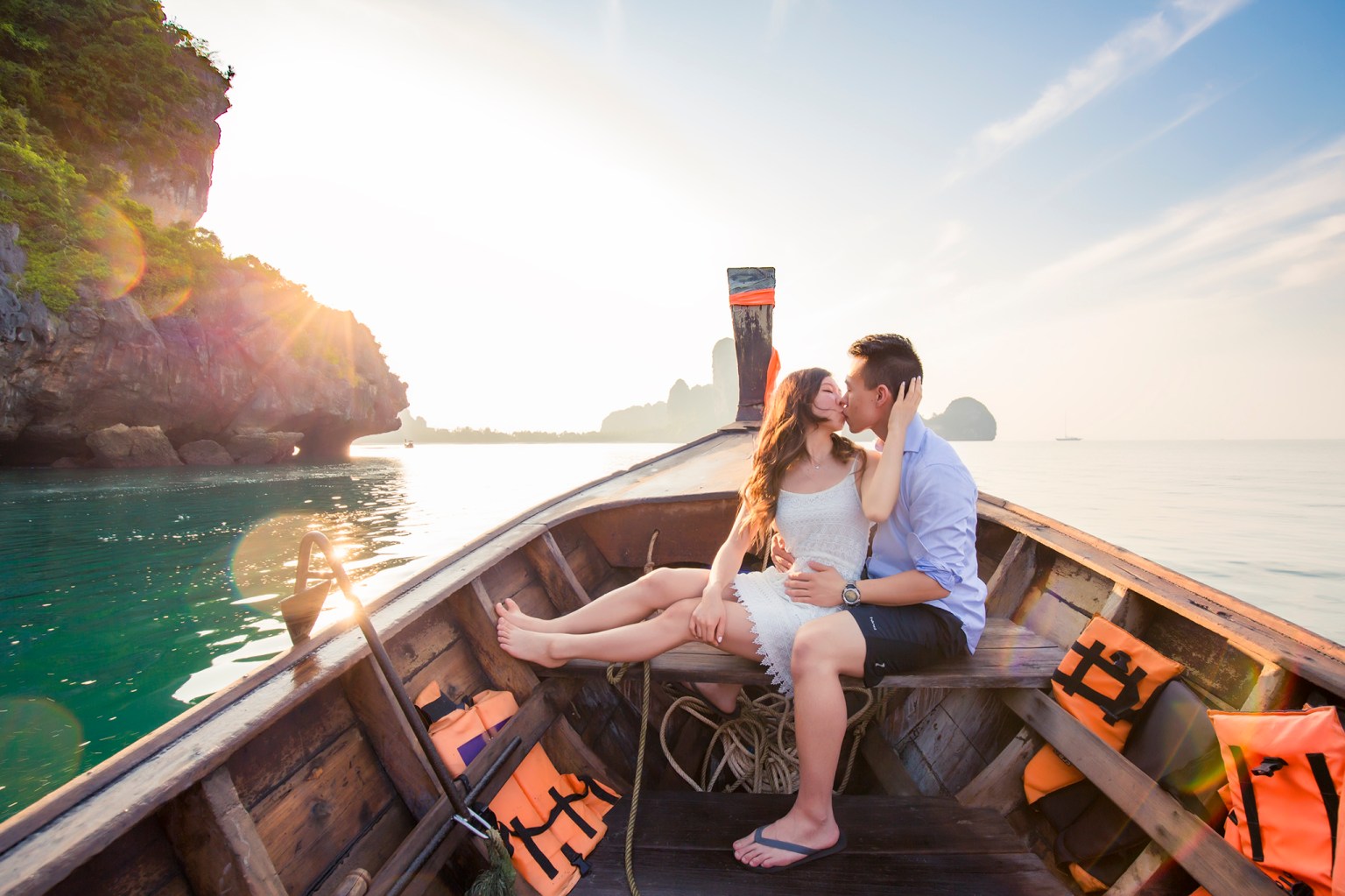 A couple sharing a romantic kiss on a boat at sunset, with scenic cliffs and clear water in the background.