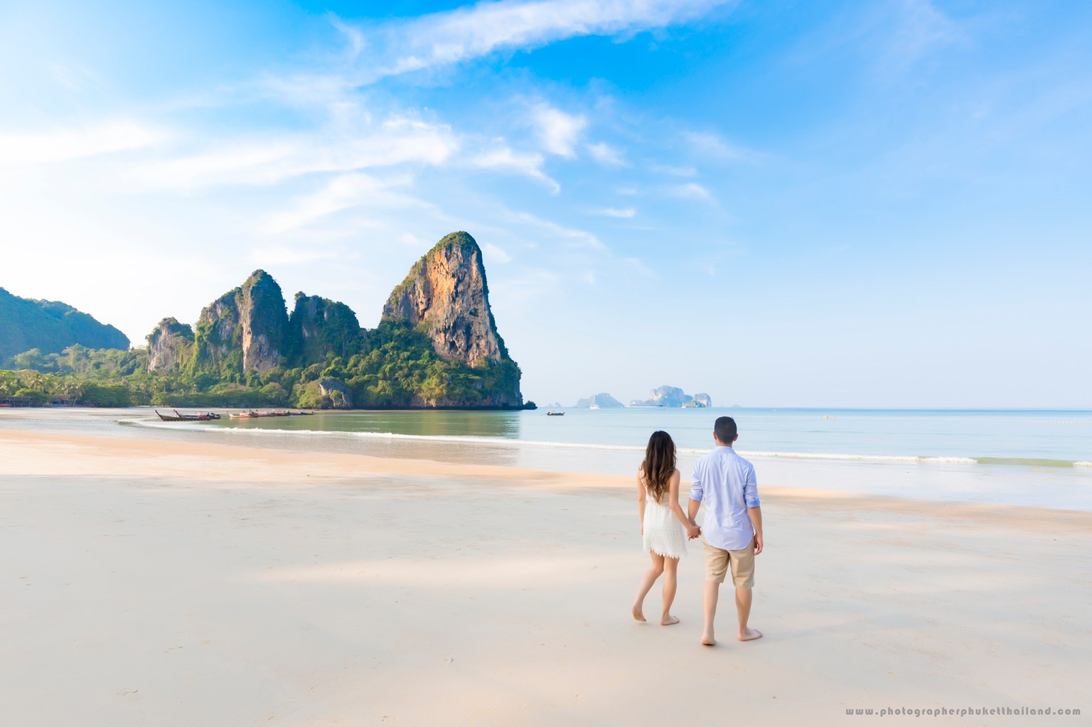 A couple walking hand in hand along a serene beach with rocky cliffs and clear blue skies in the background.