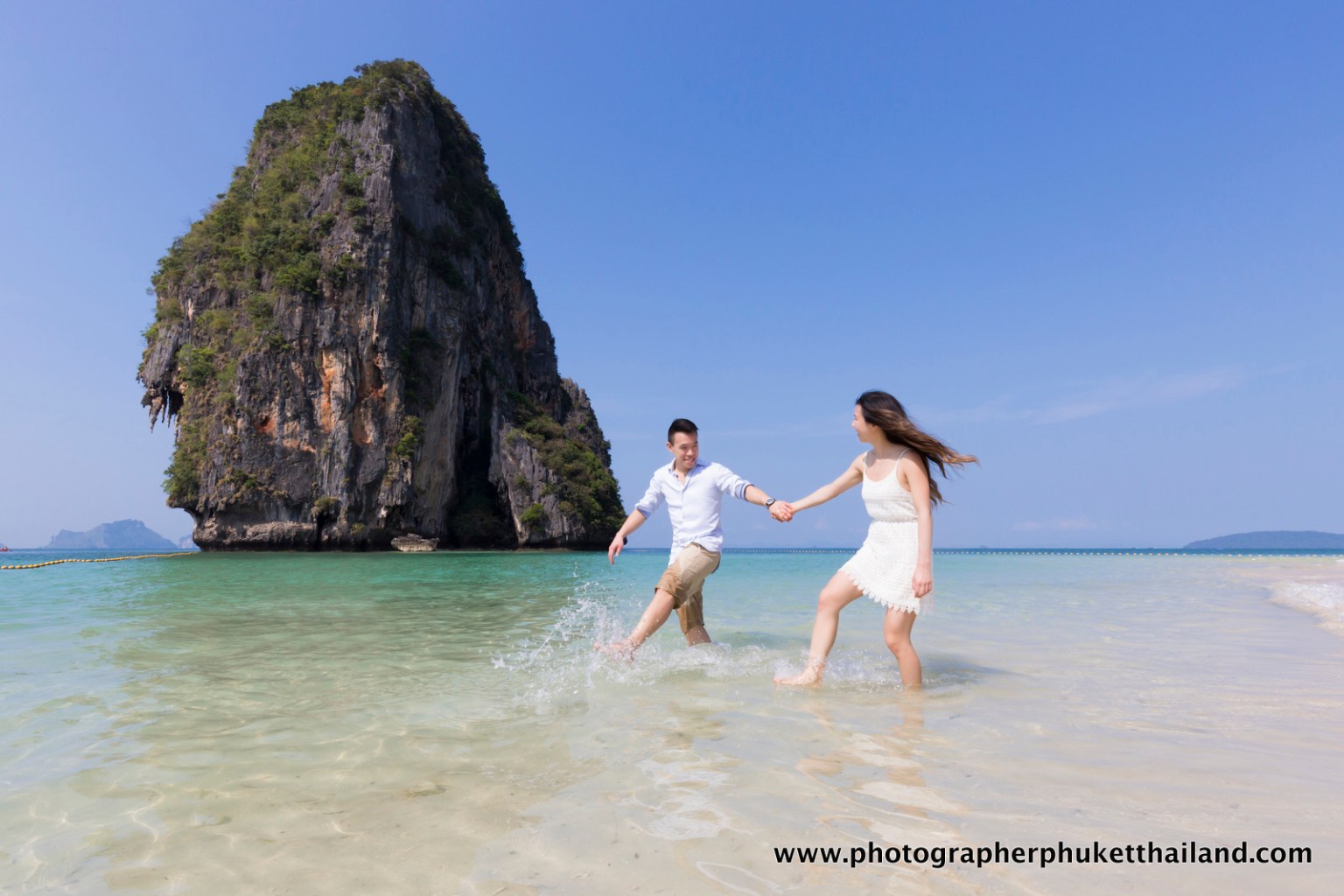 Couple playing in the shallow water on a beach in Krabi, Thailand, with a large limestone cliff in the background.