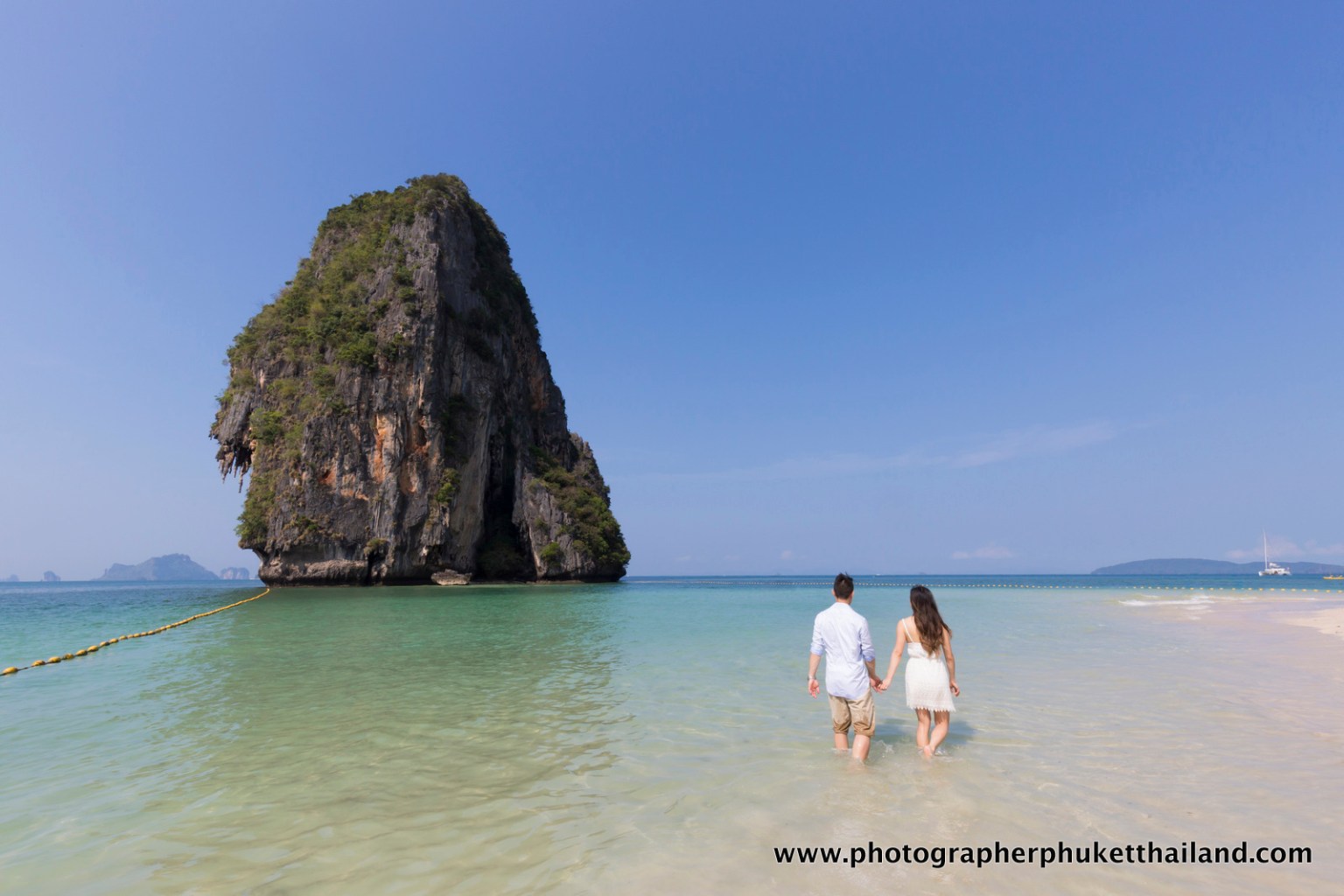 A couple holding hands while walking along a beach with a large limestone rock formation in the background and clear blue skies.