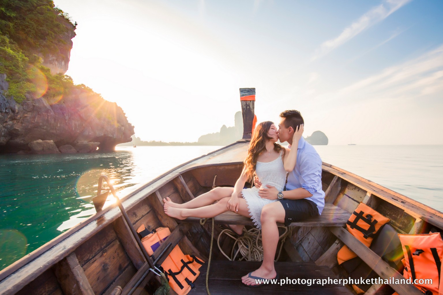 A couple kissing on a traditional boat in Krabi, Thailand, with limestone cliffs in the background and the sun setting over the water.