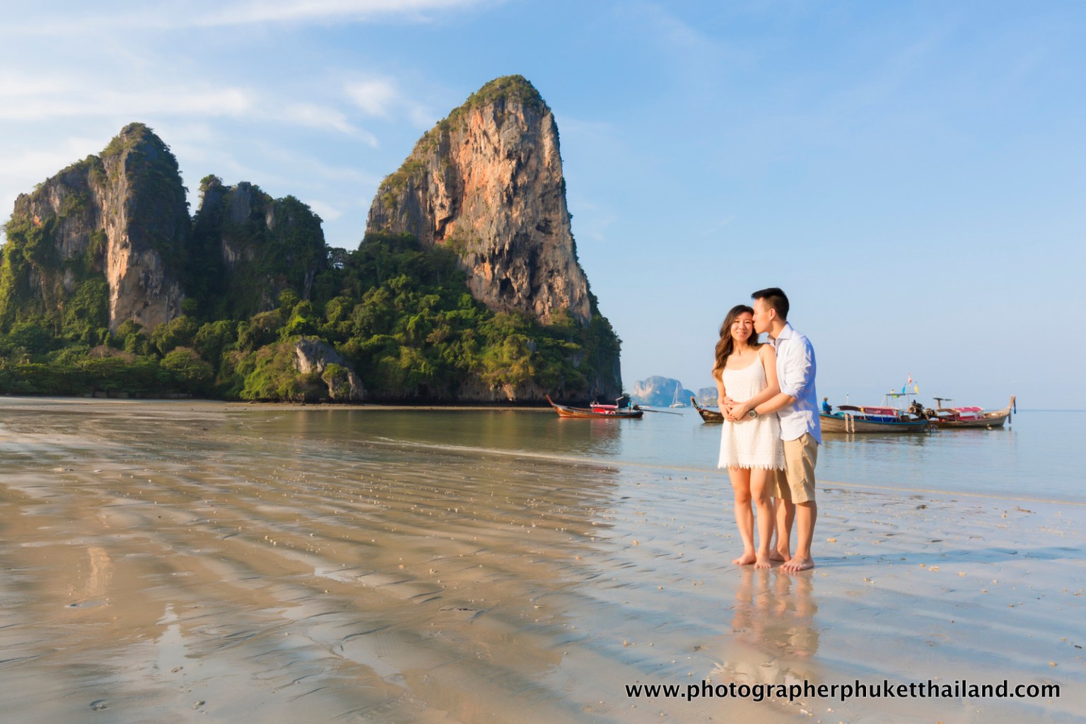 A couple standing on a railay beach, krabi smiling and embracing, with cliffs and boats in the background.